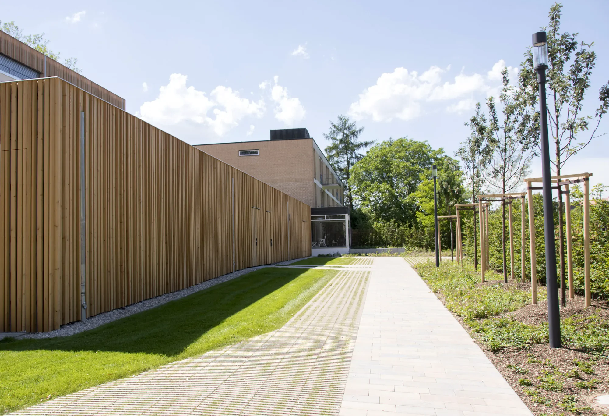 Moderner Gartenweg neben einem Holzgebäude mit Balkonen und Sitzgelegenheiten, umgeben von grünen Bäumen und Laternen bei sonnigem Himmel.
