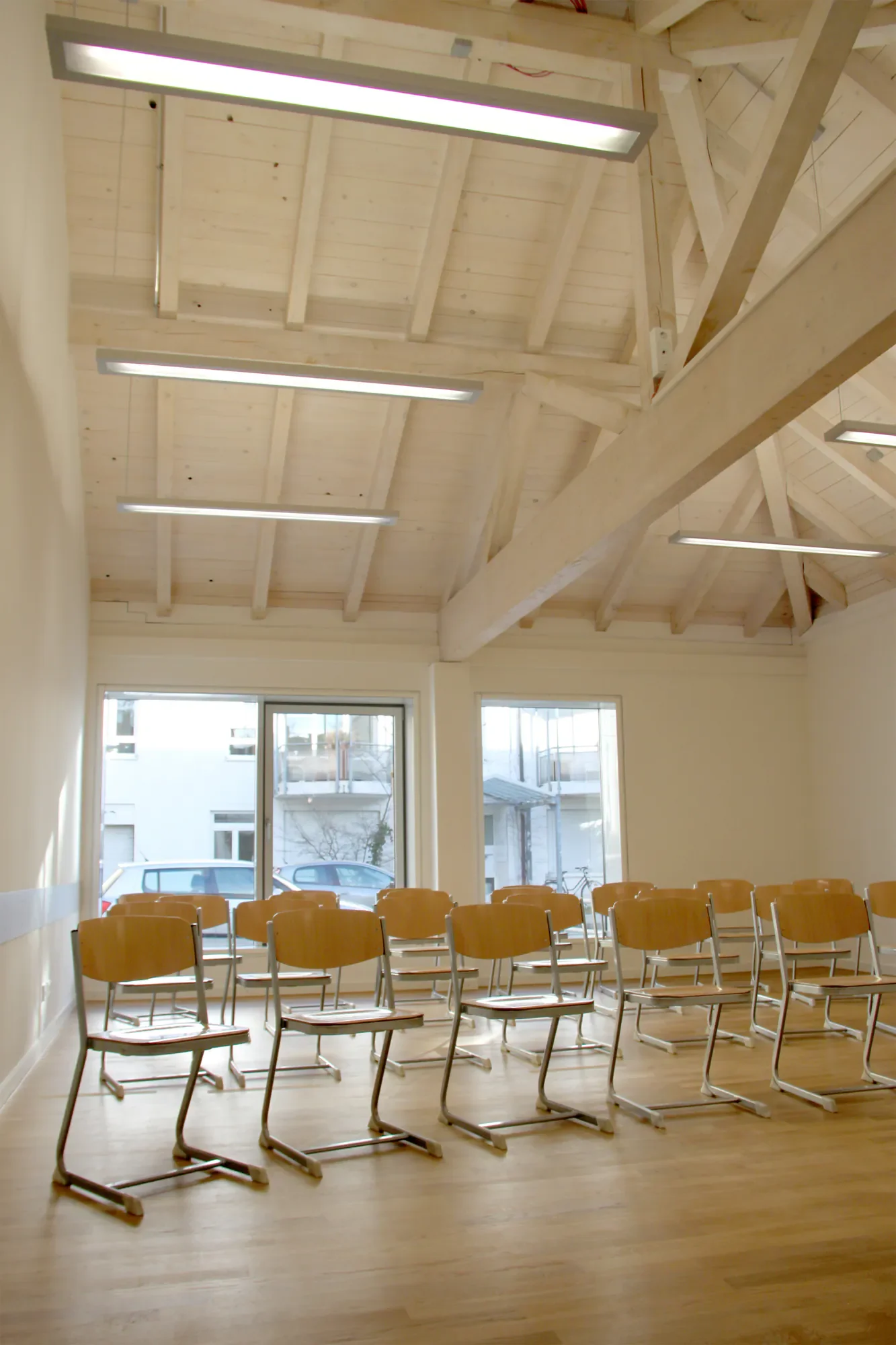Ein leerer Schulungsraum mit Reihen von Holzbänken, großen Fenstern und einer hohen Decke mit sichtbaren Holzbalken und Deckenleuchten.