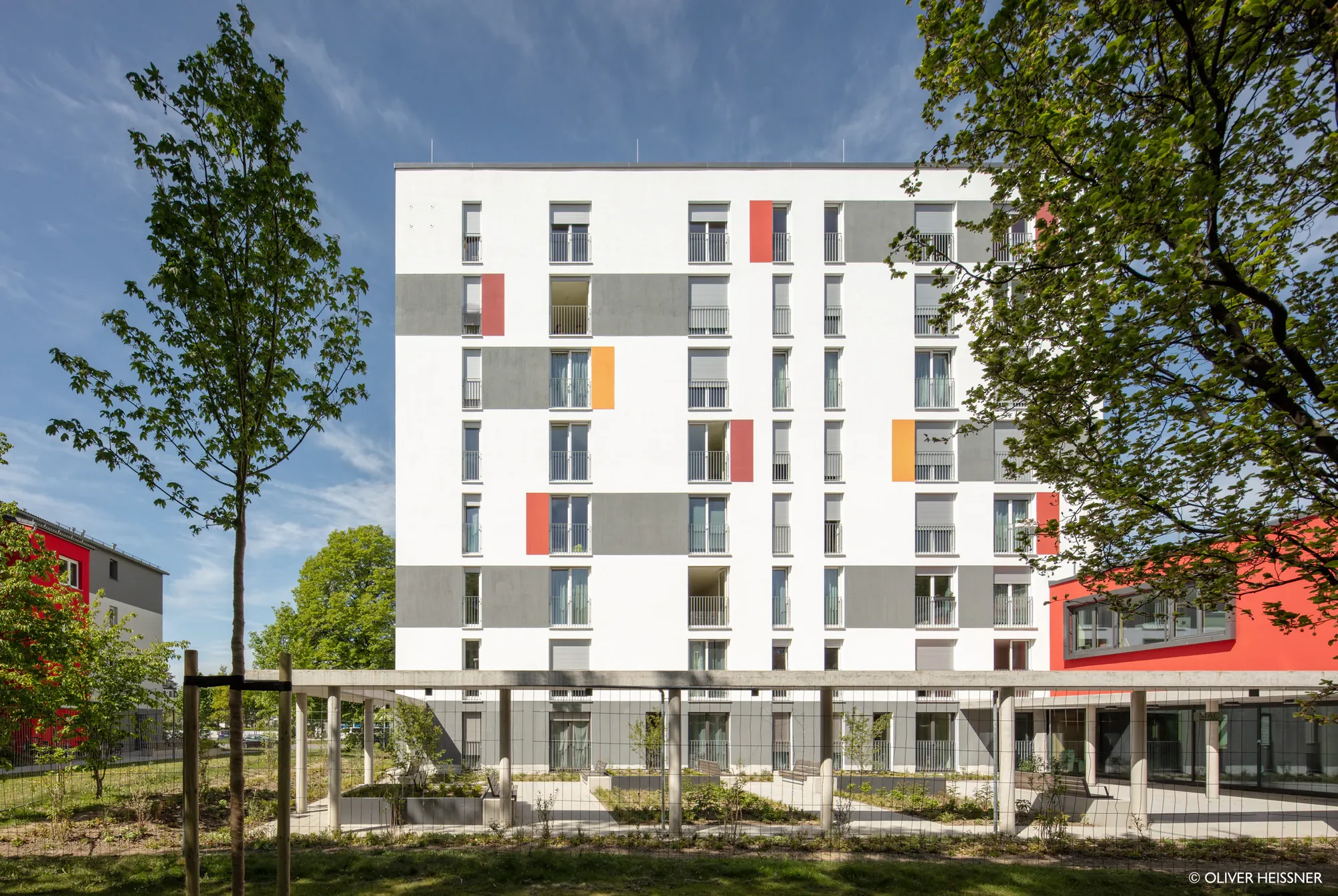 Modernes, mehrstöckiges Apartmentgebäude mit weißen, grauen und roten Akzenten, umgeben von Bäumen bei Sonnenschein.