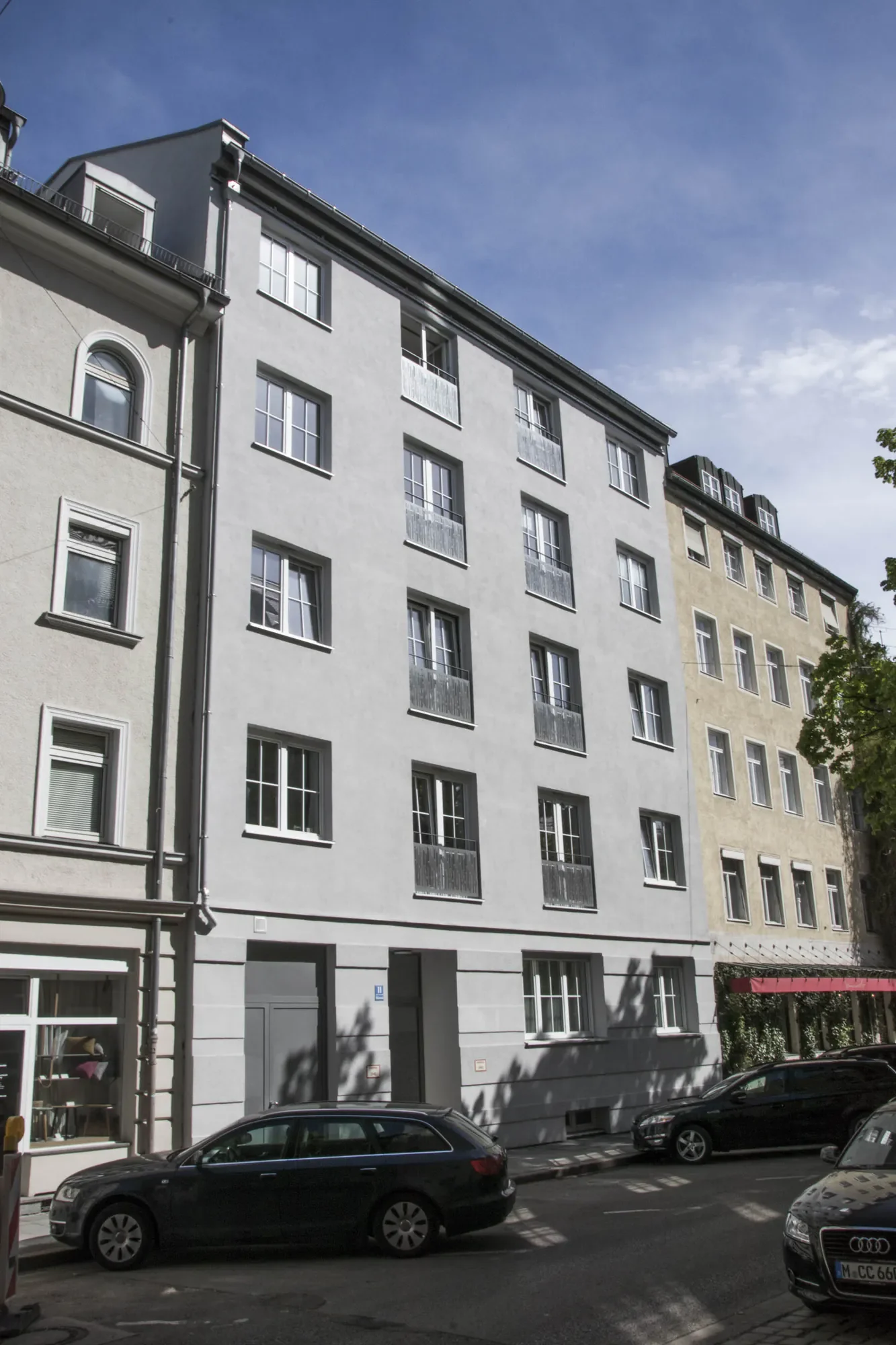 Mehrstöckiges Gebäude mit weißen Fensterrahmen und Balkonen, parkende Autos auf der Straße, einige Schatten von Bäumen, blauer Himmel mit wenigen Wolken.