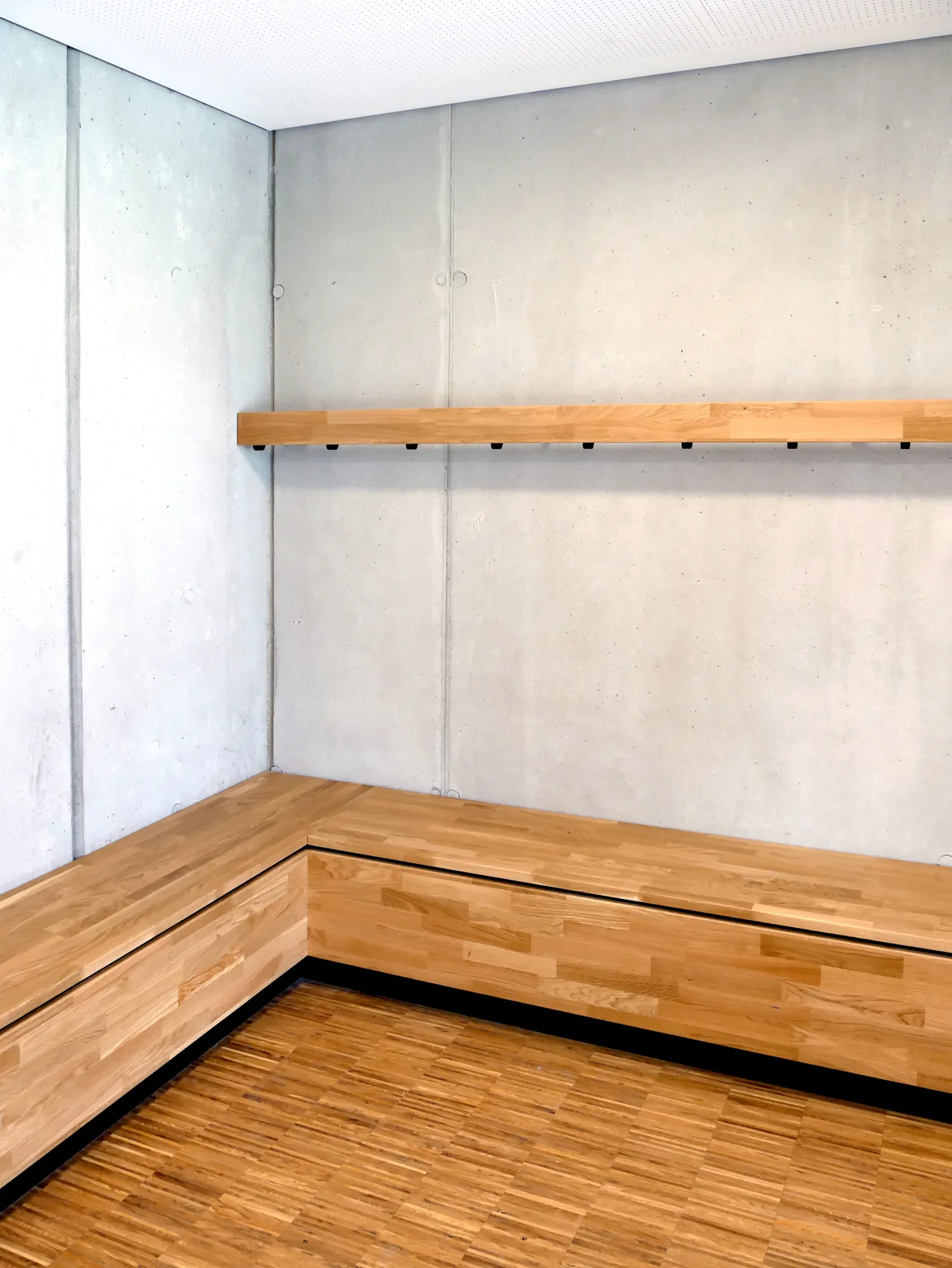 Ein Moderner Innenraum mit Holzvertäfelung, einer Betondecke und einem grau verputzten Wand. Eine Holzbank ist an der Wand befestigt.
