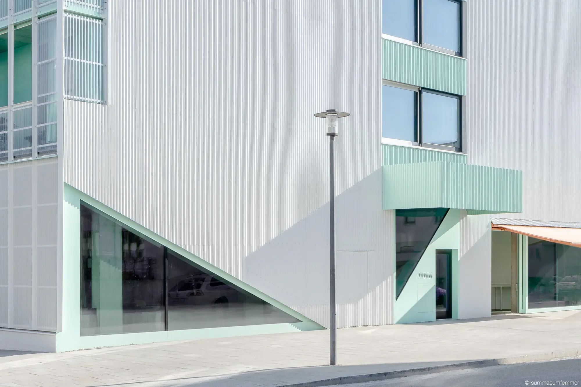 Modernes Gebäude mit weißen und grünen Fassadenelementen, großen Fenstern, einem Laternenstrahler und einer Treppe im Außenbereich.