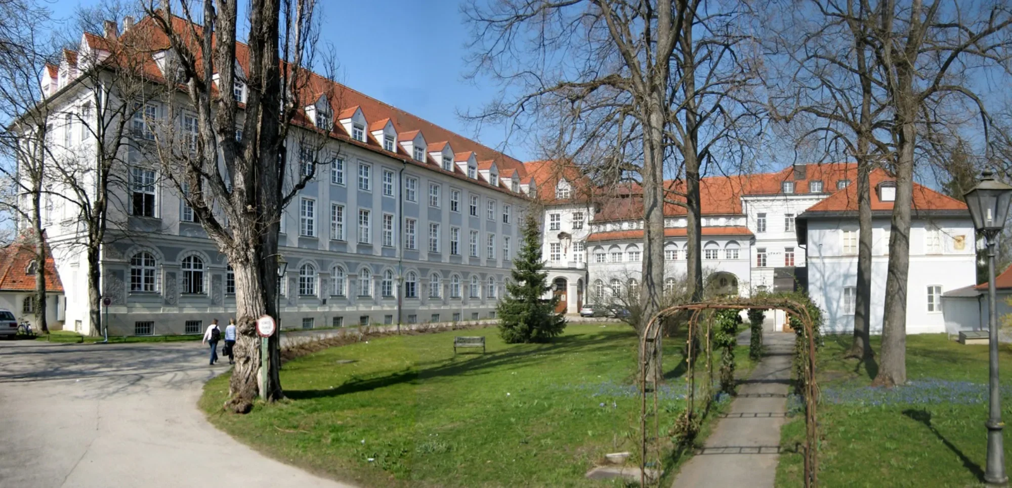 Ein großes weißes Gebäude mit rotem Satteldach, Bäumen im Vordergrund und einem kleinen Gartenweg, in einer ruhigen Umgebung bei Tageslicht.
