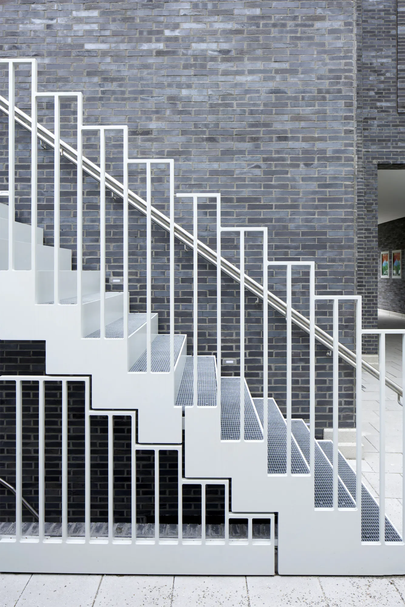 Weiße Metalltreppe vor einer grauen Ziegelmauer, mit schwarzen und weißen Galerie- und Gitterelementen im Hintergrund.