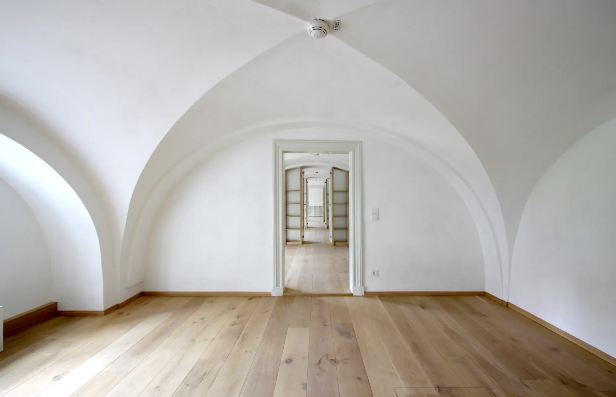 Ein heller, leerer Raum mit weißen gebogenen Decken und Holzboden, Blick durch eine Tür in einen weiteren Raum mit Holzfußboden und weißen Wänden.