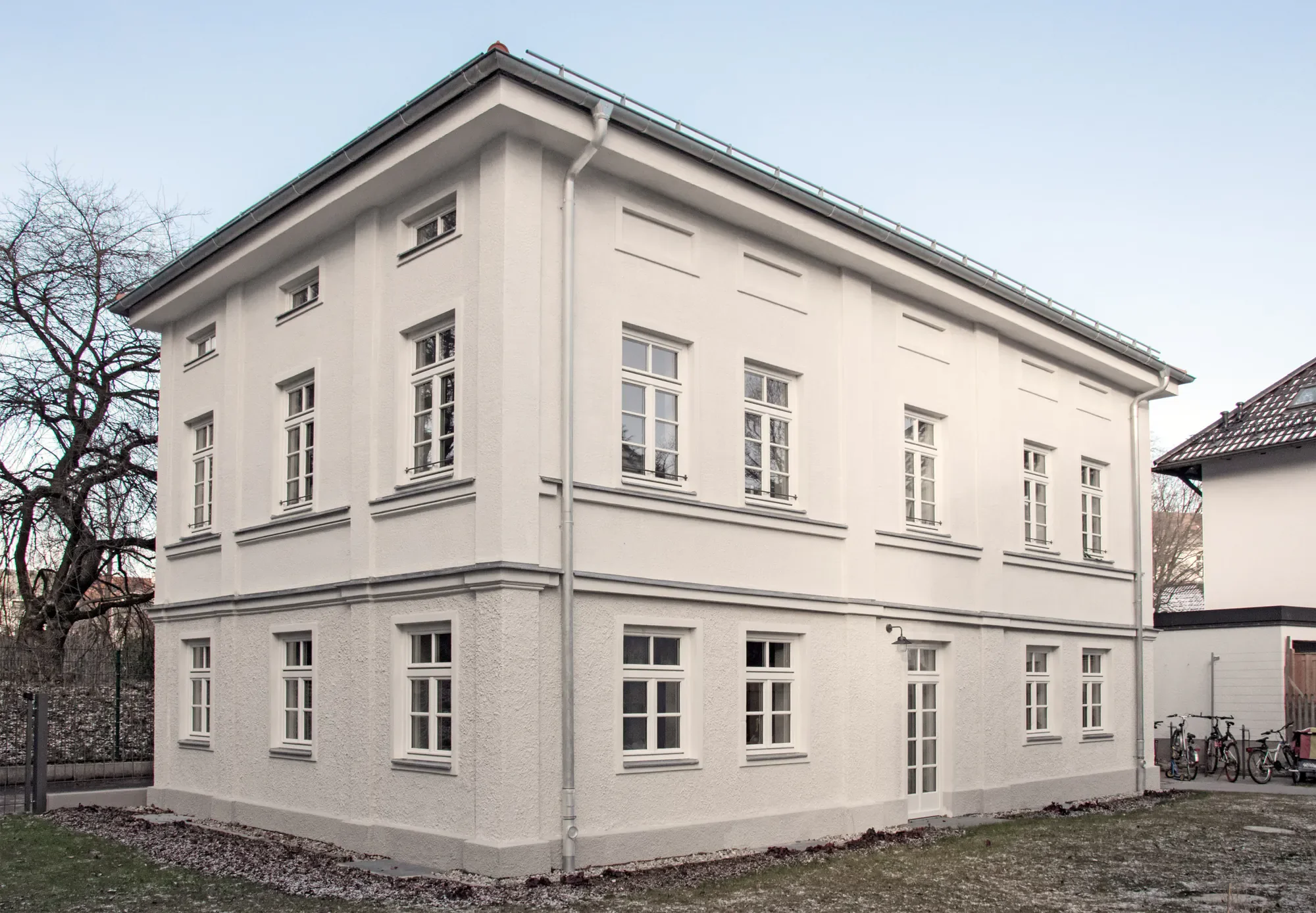 Ein weißes, mehrstöckiges Gebäude mit mehreren rechteckigen Fenstern und einer Flachdachterrasse, umgeben von Bäumen und Fahrrädern.