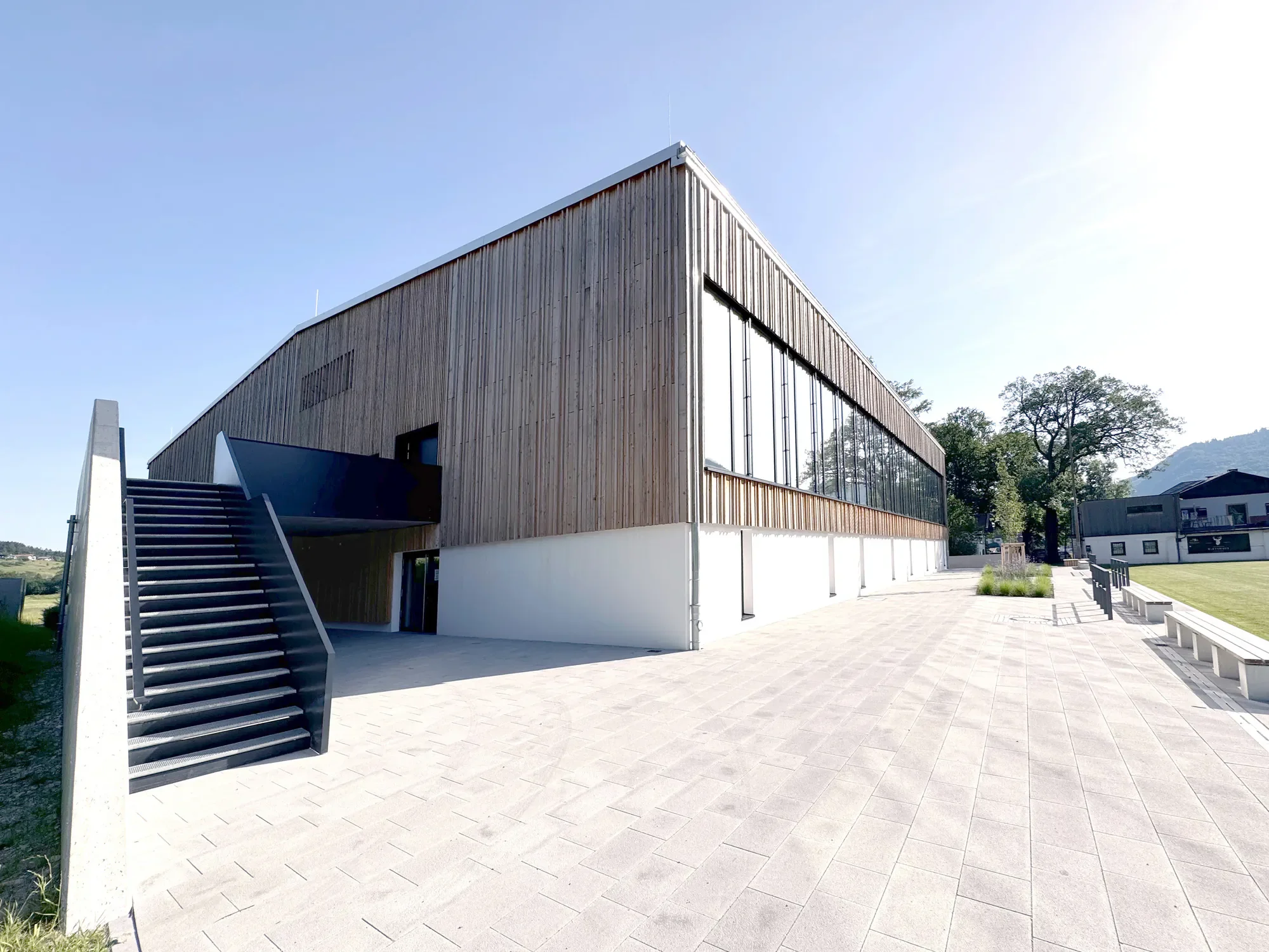 Moderner Gebäudeaussenansicht mit Holzfassade und großen Fenstern, bei sonnigem Himmel, umliegende Bäume, gepflasterte Fläche mit Sitzbänken.