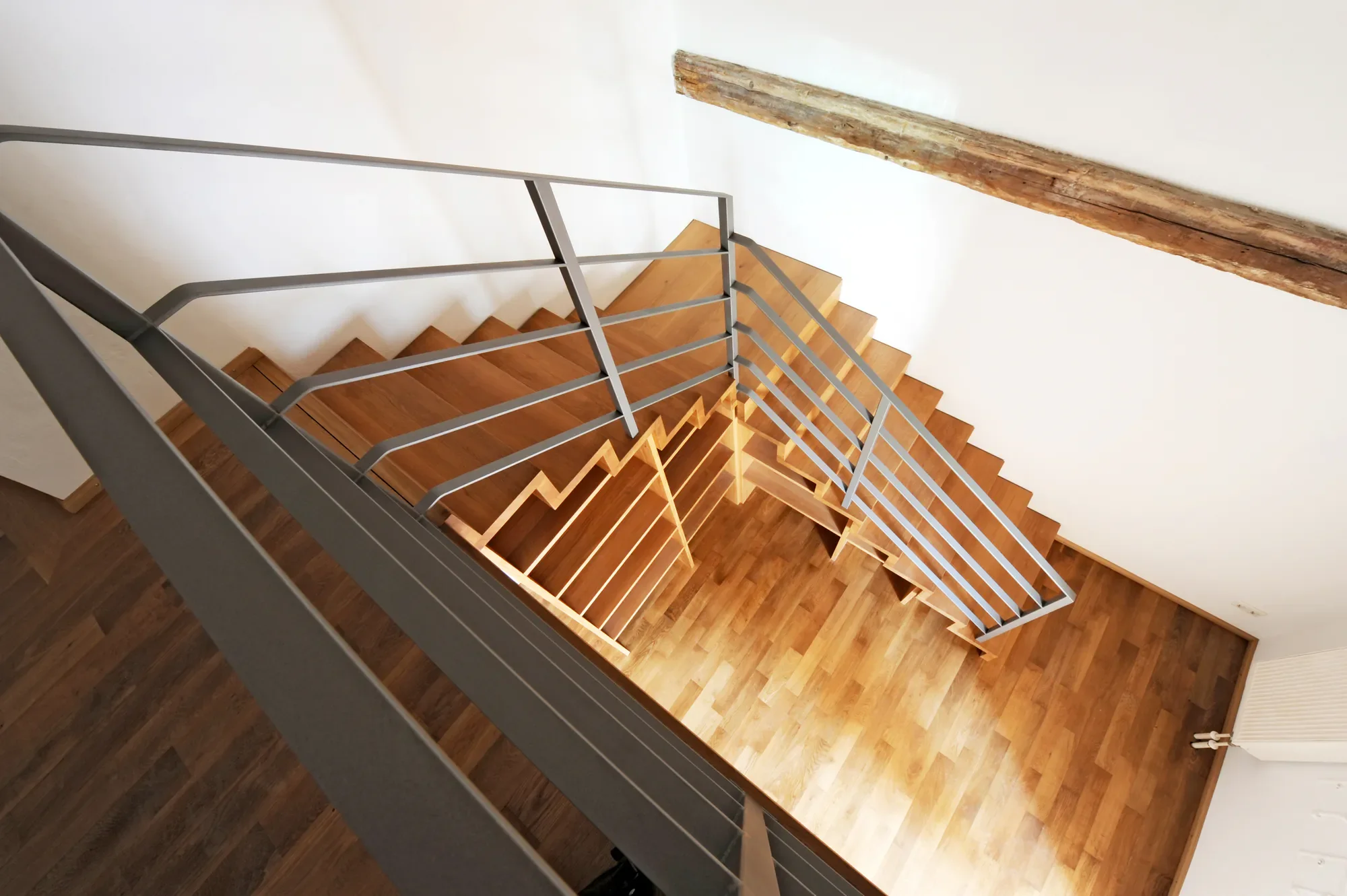 Heller Treppenaufgang mit Holzstufen, grauem Handlauf und einem alten Holzbalken an der Wand, Blick nach oben.