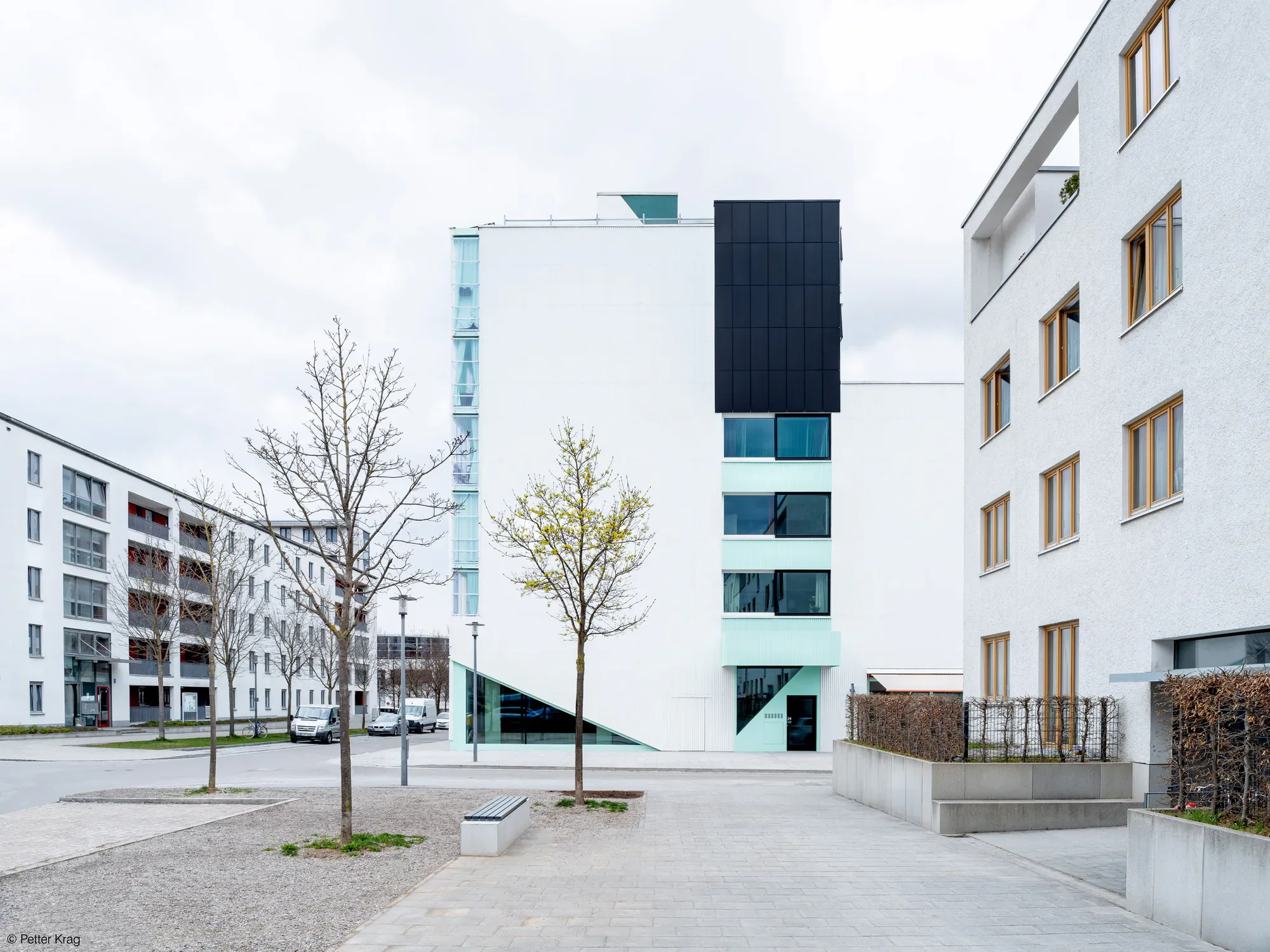 Moderne weiße Wohngebäude mit großen Fenstern und Balkonen in einem urbanen Gebiet, Bäume im Vordergrund, bewölkter Himmel.