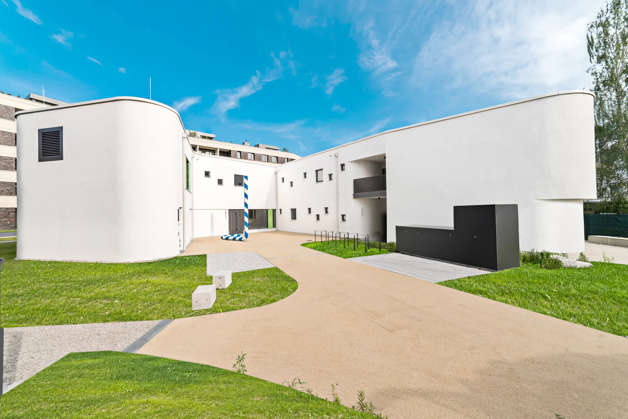 Modern weißes Gebäude mit glatten Wänden, kleinen Fenstern und einer schwarzen Tür, umgeben von grünem Rasen und einem hellen Gehweg, unter blauem Himmel mit einigen Wolken.