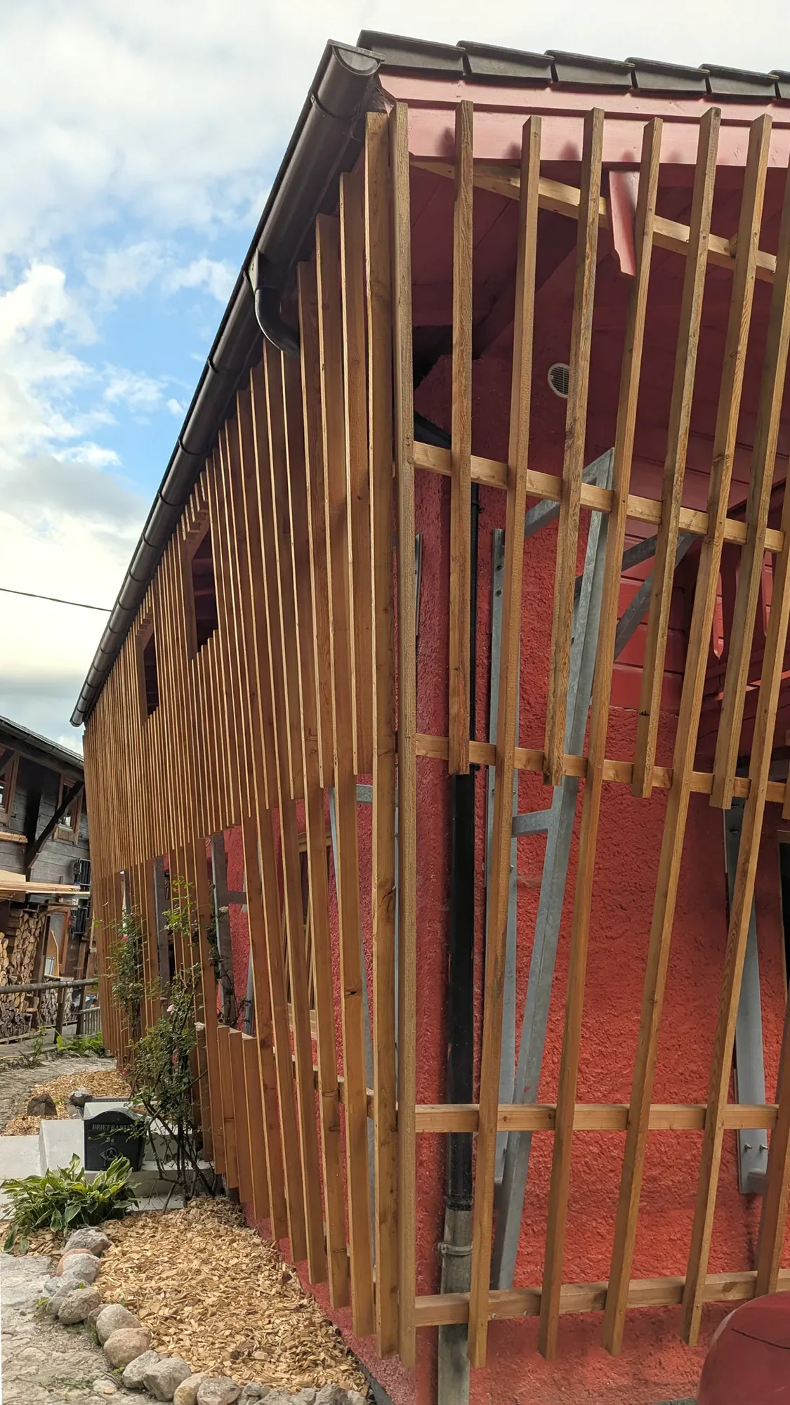 Der Bau eines Holzrahmen für ein Gebäude, mit vertikalen Holzlatten an einer roten Wand, eine Leiter im Hintergrund, unter einem Dach.