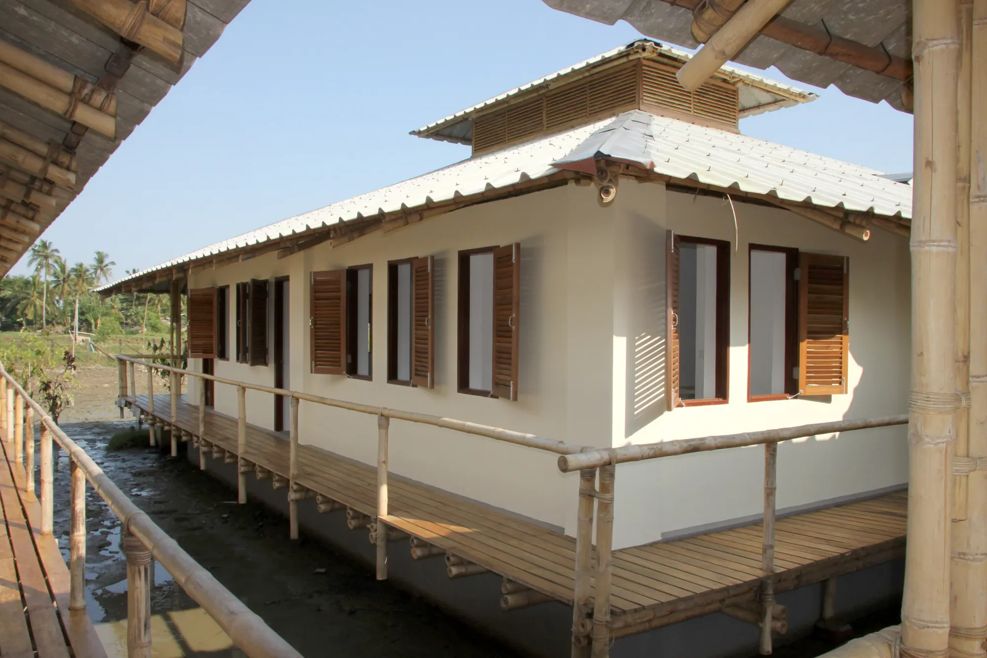 Ein Haus mit weißen Wänden, Holztüren und -fenstern, auf Stelzen gebaut, mit einer Holzveranda. Das Haus befindet sich in einer ländlichen Umgebung mit Palmen im Hintergrund. Es ist sonnig und der Himmel ist klar.