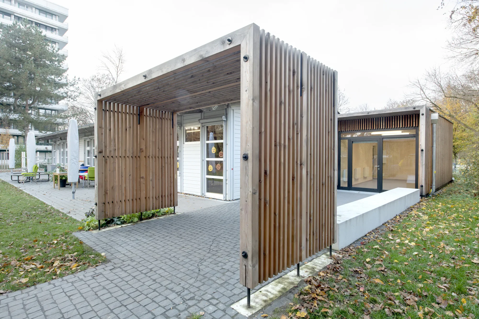 Moderne Holz-Kabine mit überdachtem Eingangsbereich und Glastür, umgeben von Bäumen und einer gepflasterten Terrasse im Freien.