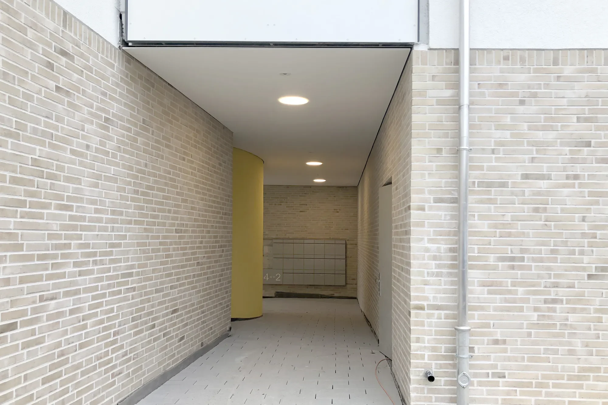 Ein Durchgang zwischen zwei weiß verputzten, gemauerten Gebäuden mit Ziegelfassade. Im Hintergrund sind Briefkästen und nummerierte Wandtafeln sichtbar, in der Decke sind eingelassene Beleuchtungsstrahler installiert.