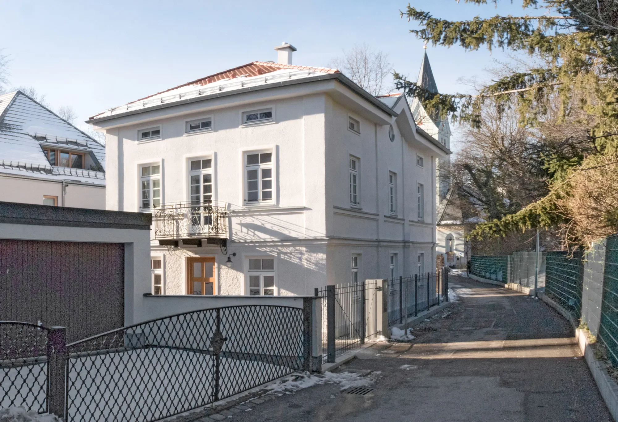 Ein weißes mehrstöckiges Haus mit mehreren Fenstern und Balkon, umgeben von Zäunen und Bäumen im Winter.