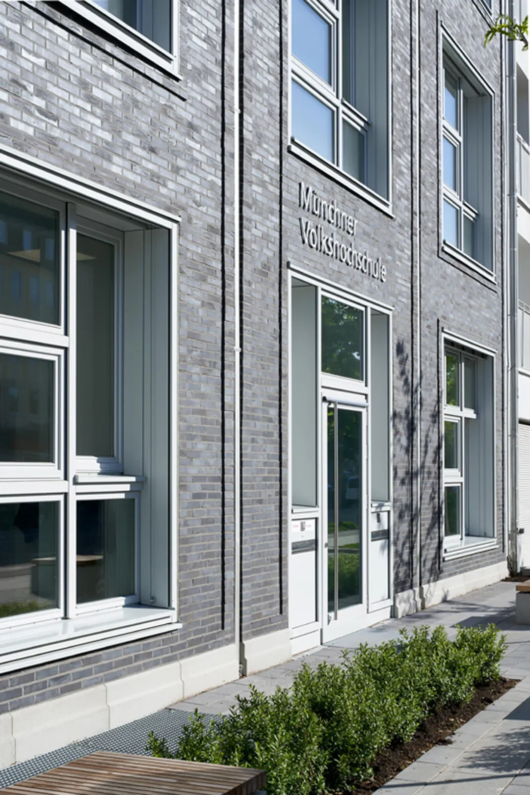 Das Bild zeigt die Fassade eines modernen Gebäudes mit grauen Ziegelsteinen und weißen Fensterrahmen. Es trägt das Schild 'Münchner Volkshochschule'.