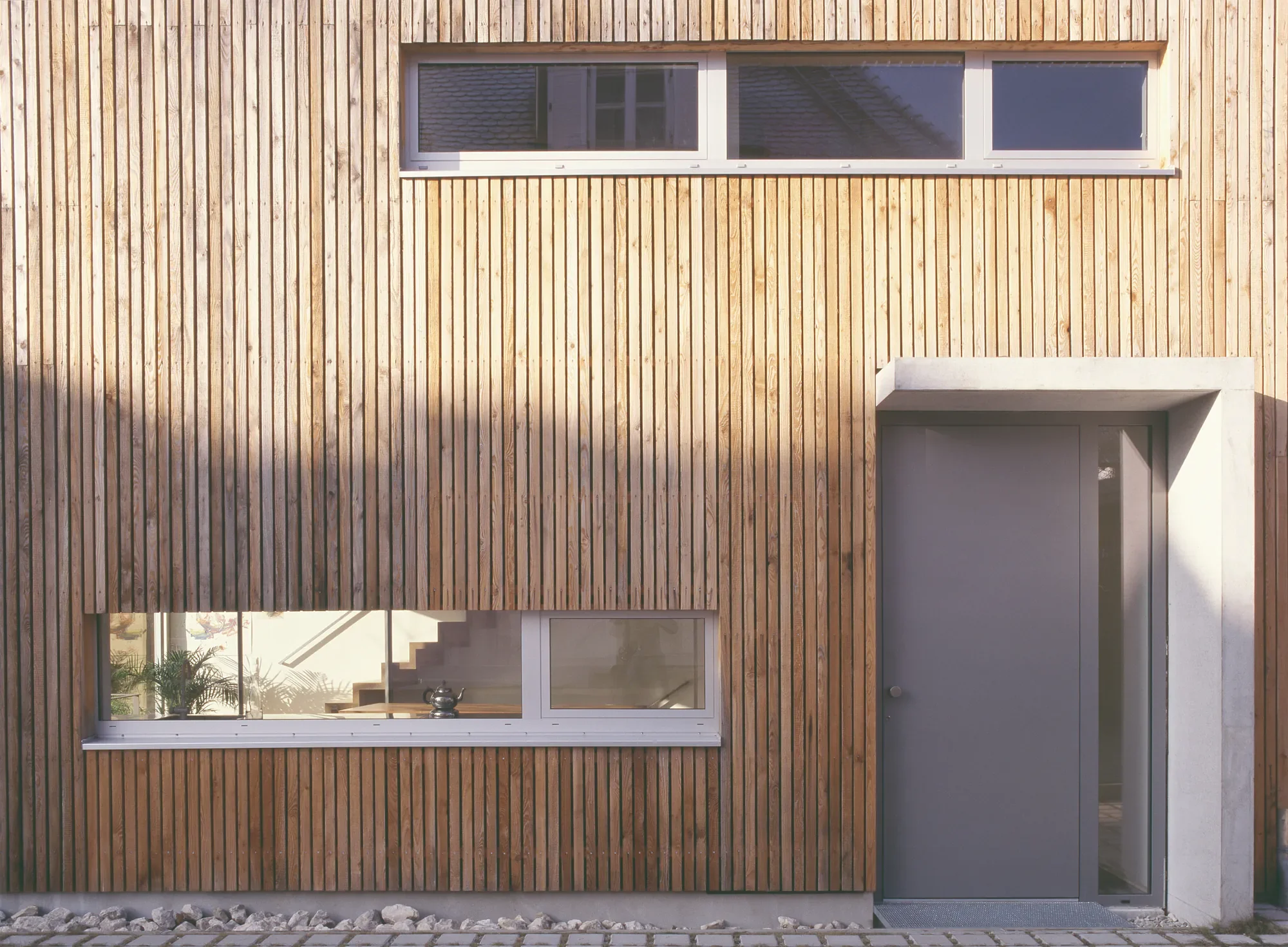 Moderne Hausfassade mit Holzvertäfelung, großes Fenster, grauem Eingangstür und niedrigem weißen Anbauteil.