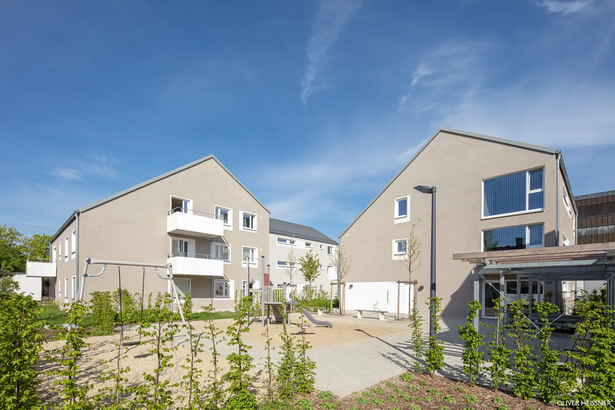 Moderne mehrstöckige weiße Wohnhäuser mit großen Fenstern, Balkonen und einem Spielplatz im Vordergrund, unter einem blauen Himmel mit wenigen Wolken.