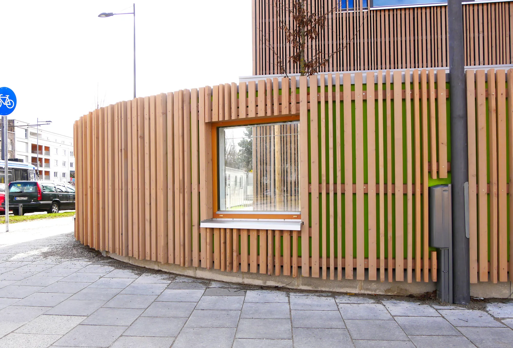 Modernes Gebäude mit Holzvertäfelung, Fenster und kleinen Baum auf Dach, an einer Straßenkreuzung in einer urbanen Umgebung.