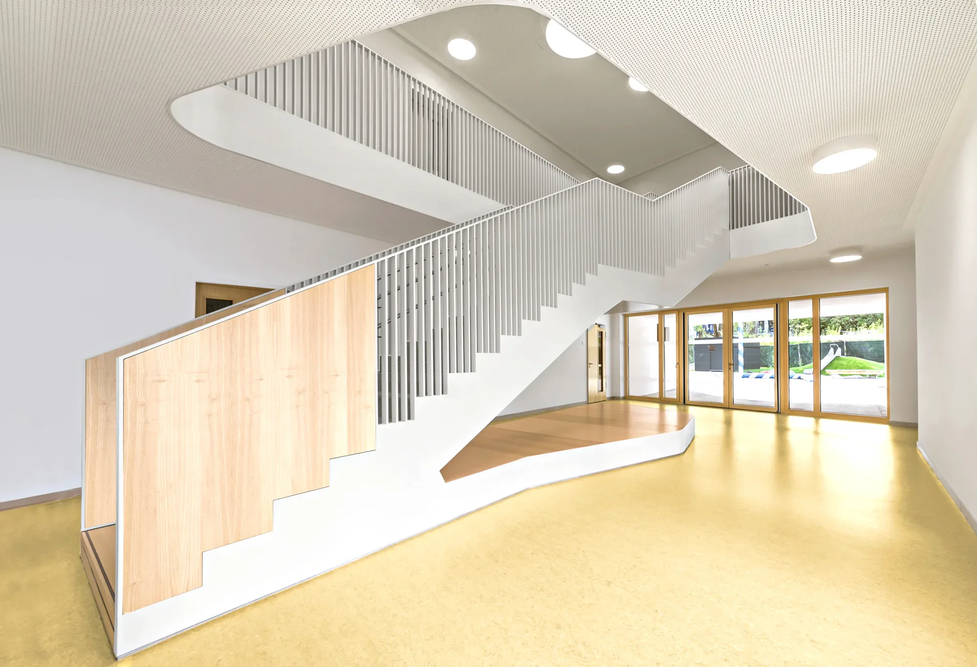 Heller Raum mit Treppe, große Fenster, gelber Boden, weiße Wände, Holz- und Metallgeländer, helle Decke mit runden Lichtstrahlern.