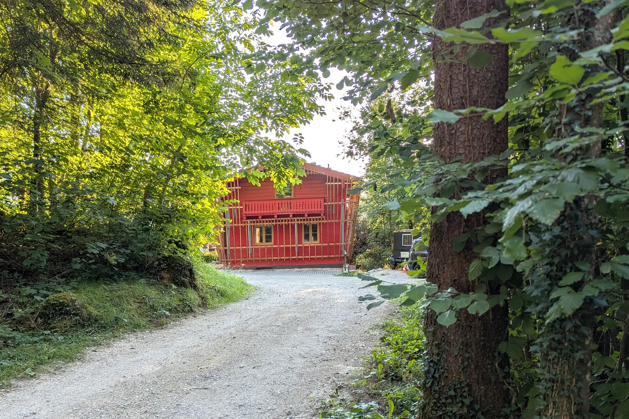 Ein rotes Haus im Bau, umgeben von grünen Bäumen und Sträuchern, auf einer Kiesstraße in einem bewaldeten Gebiet.