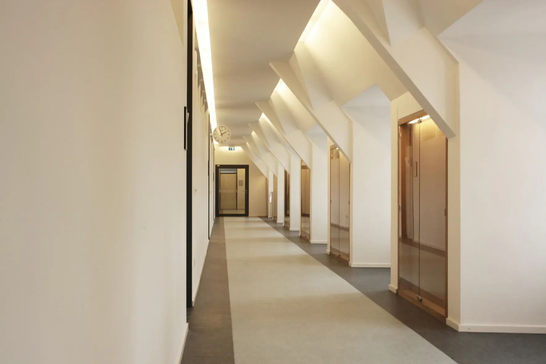 Gerade Flur in einem modernen Gebäude mit weißen Wänden, Deckenleuchten, Aufzügen und einem Wandwecker, Blick in die Tiefe.
