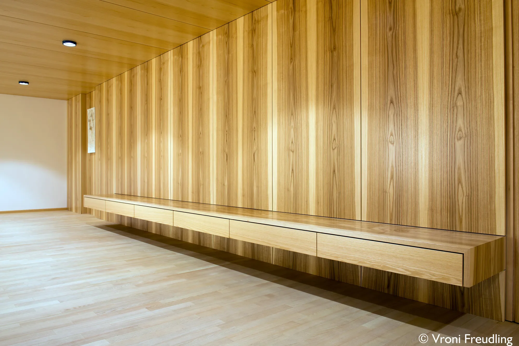 Innenraum mit holzverkleideten Wänden und Decke, Sitzbank aus Holz, zwei Deckenspots, helles Holzboden.