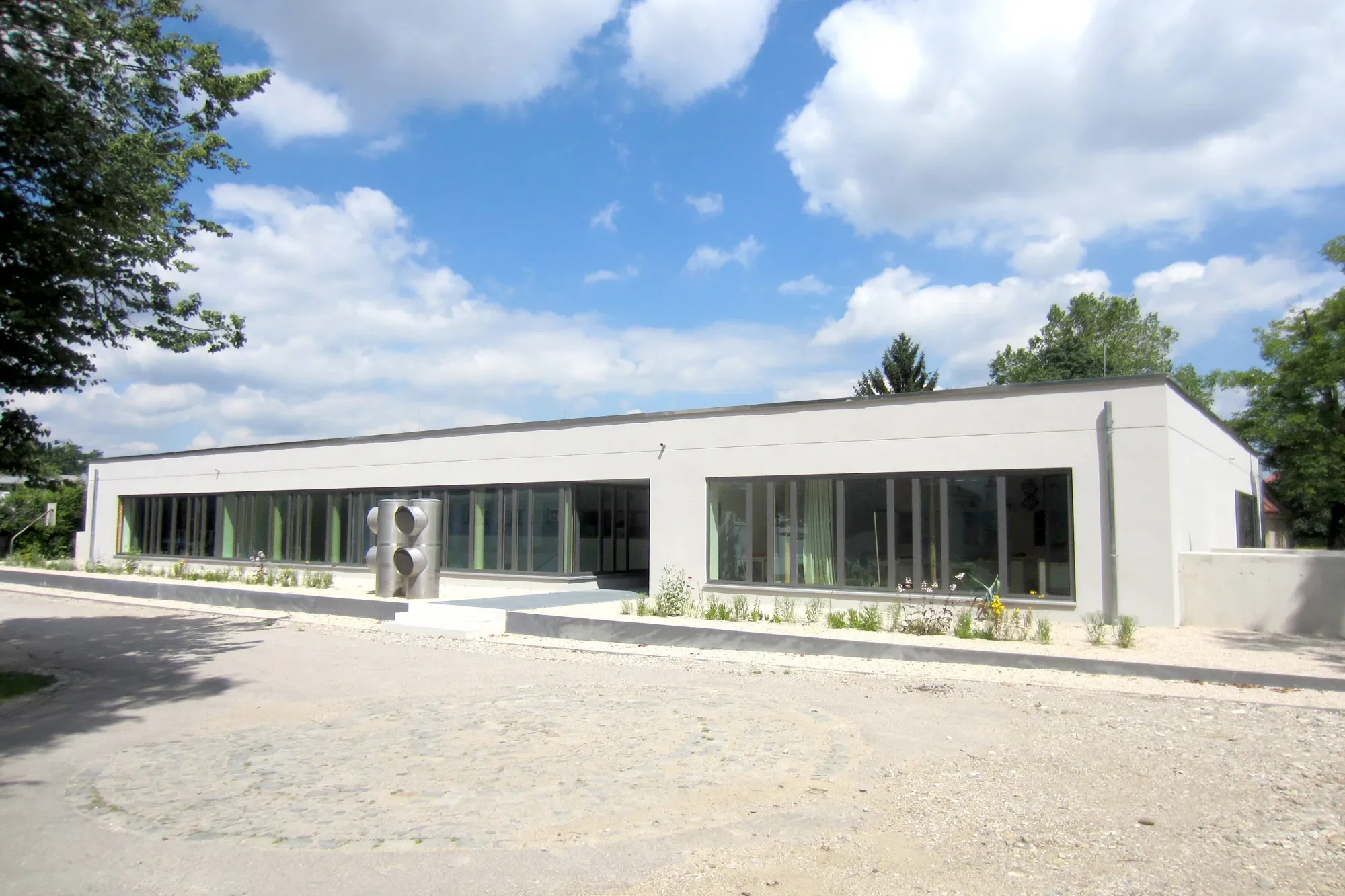 Ein modernes weißes Gebäude mit großen Fenstern, umgeben von Bäumen und einem blauen Himmel mit Wolken, im Freien.