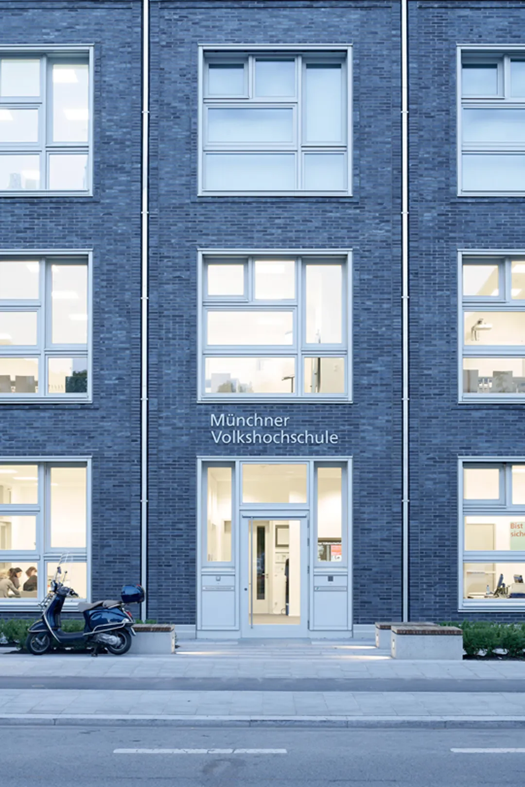Das Bild zeigt das Gebäude der Münchner Volkshochschule, eine moderne bläuliche Backsteinfassade mit mehreren Fenstern und einem Eingang. Vor dem Gebäude ist eine Straße mit einem geparkten Scooter.