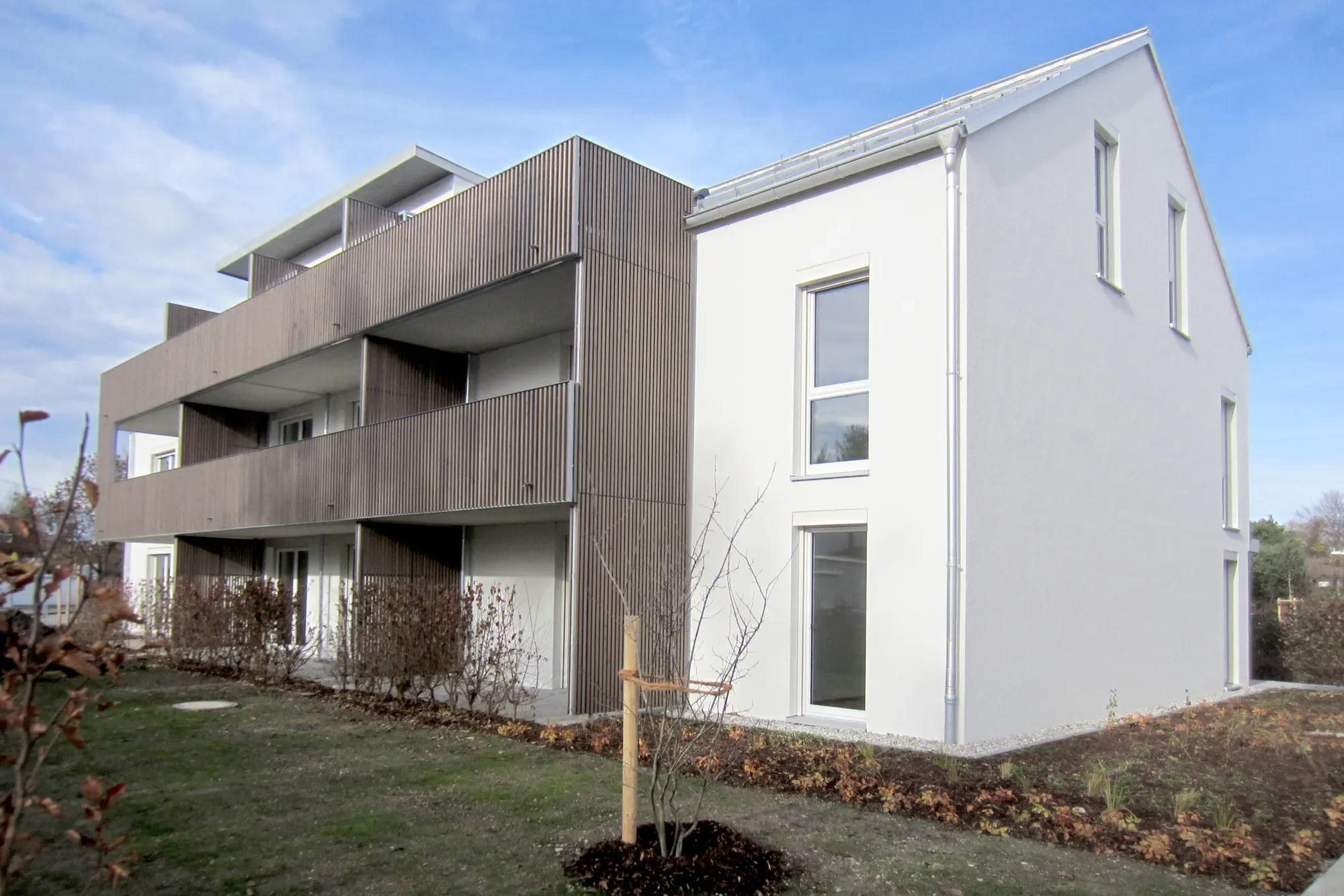 Modernes Mehrfamilienhaus mit weißen Wänden, Holzverkleidungen an der Balkonseite und großen Fenstern, umgeben von einer gepflegten Grünfläche mit kleinen Bäumen und Sträuchern bei sonnigem Himmel.