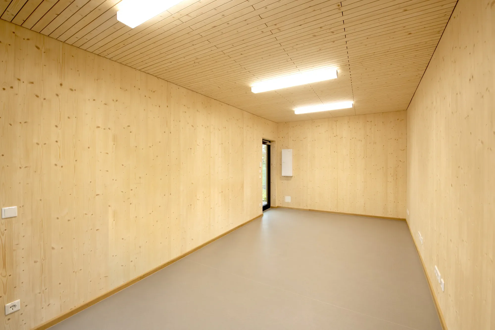 Innenraum eines Raumes mit Holzwänden und -decke, runder Deckenleuchte, Fenster und weißen Steckdosen.