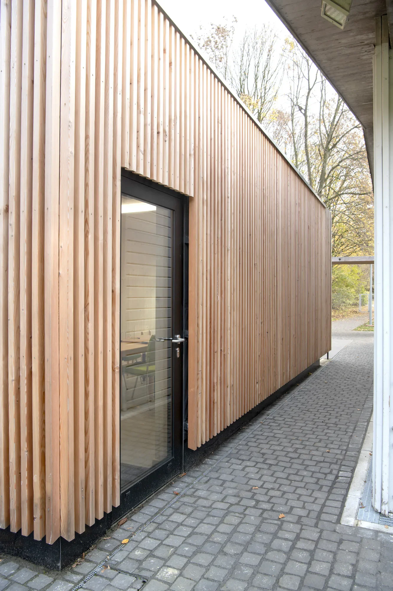 Außenansicht eines modernen Gebäudes mit Holzverkleidung, Glastür und Ziegelpflaster auf dem Boden, im Hintergrund Bäume