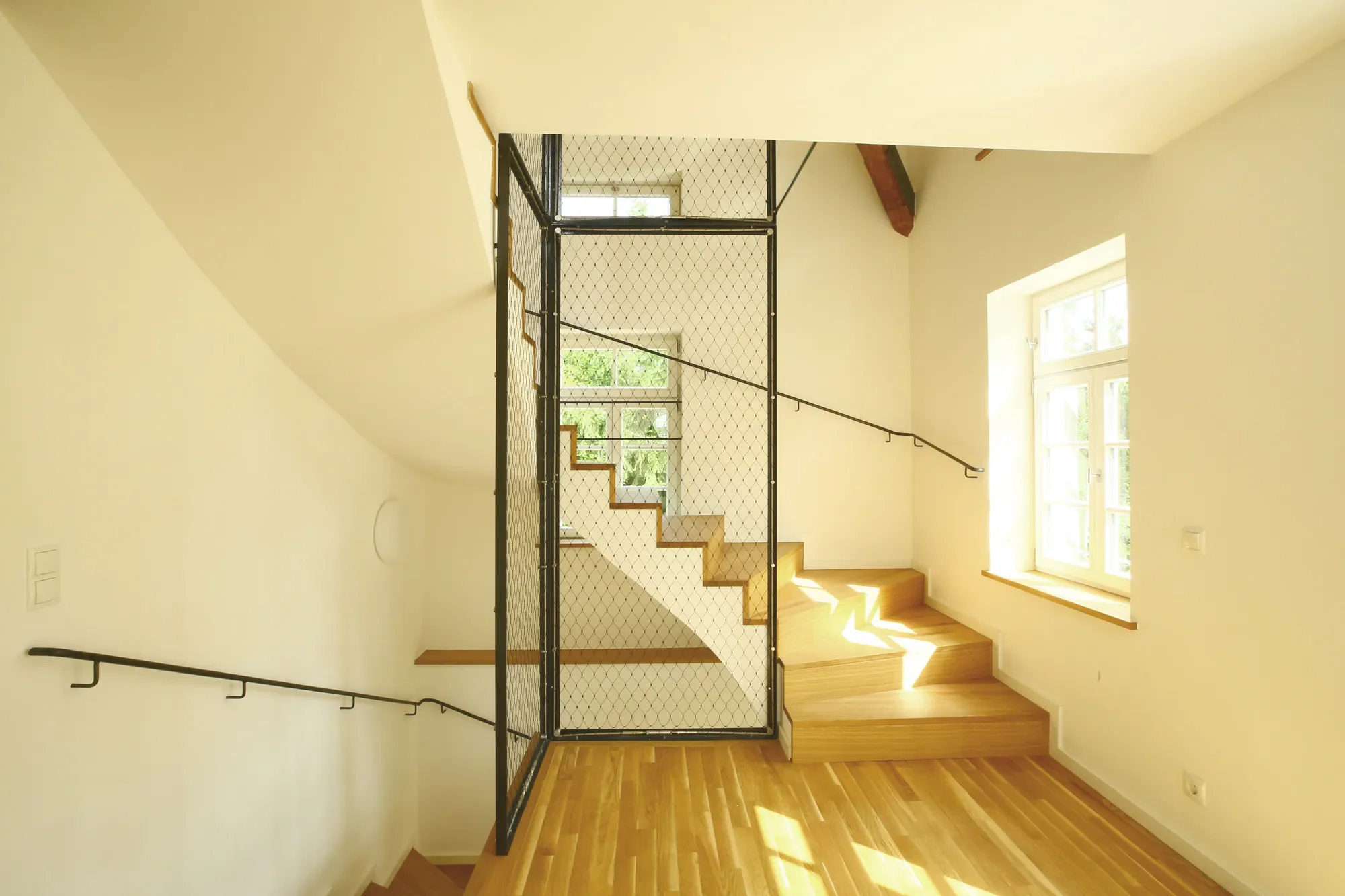 Ein heller Raum mit großen Fenstern und Holzfußboden, der eine Treppe mit Geländer und einem Sicherheitsnetz zeigt.