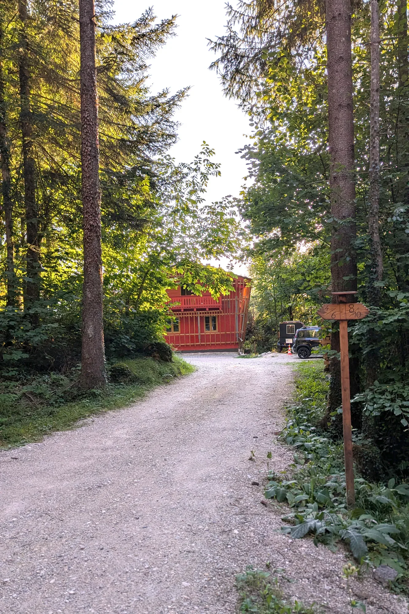 Weg durch den Wald, links und rechts Bäume, im Hintergrund ein rotes Gebäude, auf dem Weg stehen Fahrzeuge, inklusive ein schwarz-graues Auto. Es ist Tag, das Sonnenlicht scheint durch die Bäume, und es gibt einen Wegweiser auf der rechten Seite des Pfades.