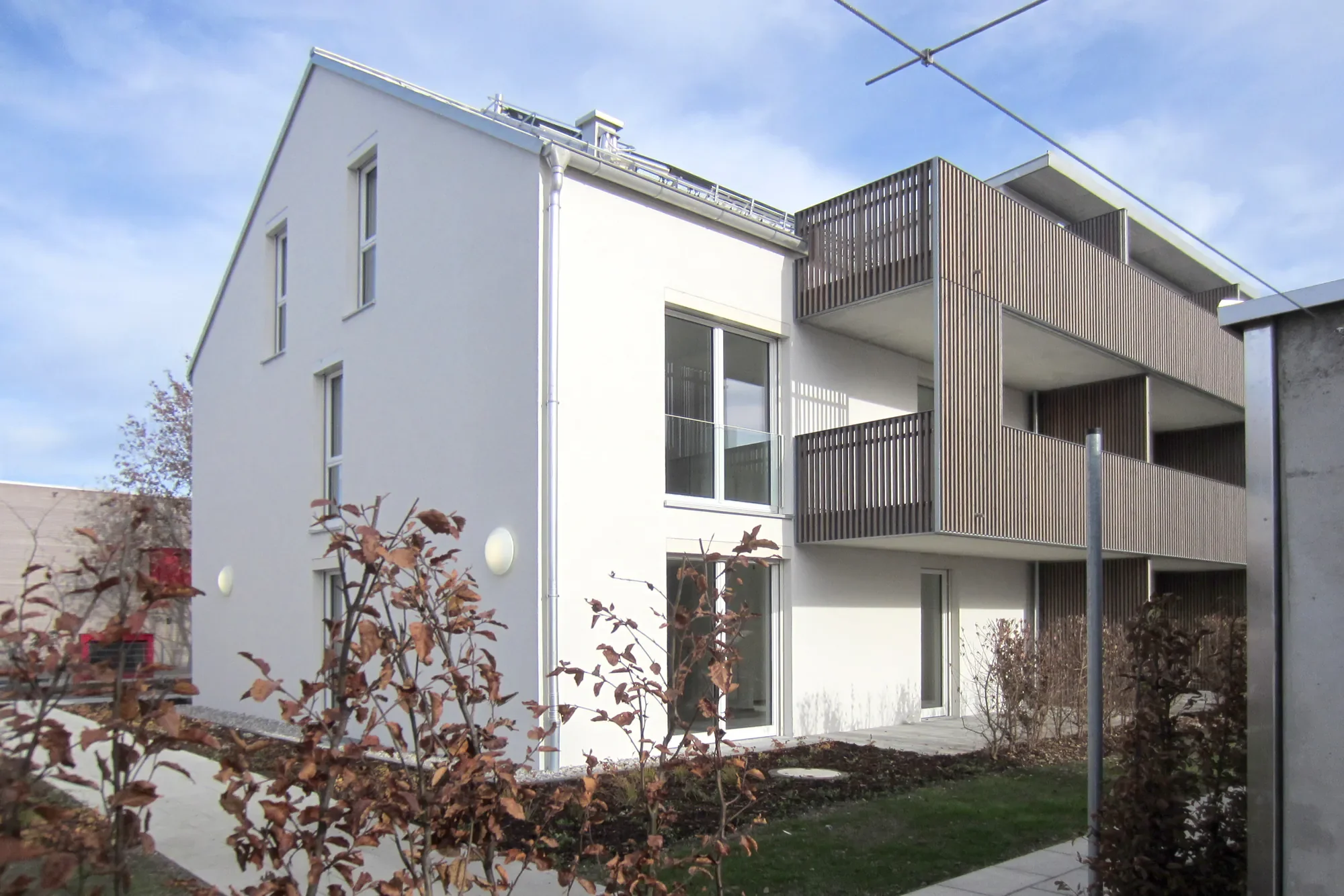 Modernes weißes Mehrfamilienhaus mit Holzverkleidungen an den Balkonen, große Fenster, Balkon mit Glasbrüstung, umgeben von Garten und Sträuchern, bei sonnigem Himmel.