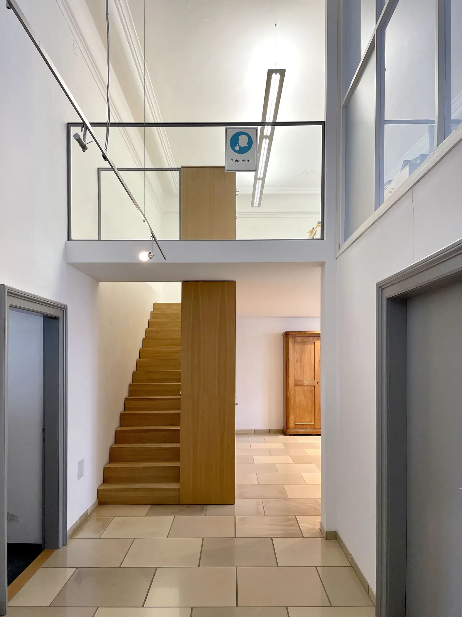 Innenansicht eines Gebäudes mit Treppe, Flur und Aufzügen, im oberen Bereich ein Schild mit der Aufschrift "Ruhe bitte!"