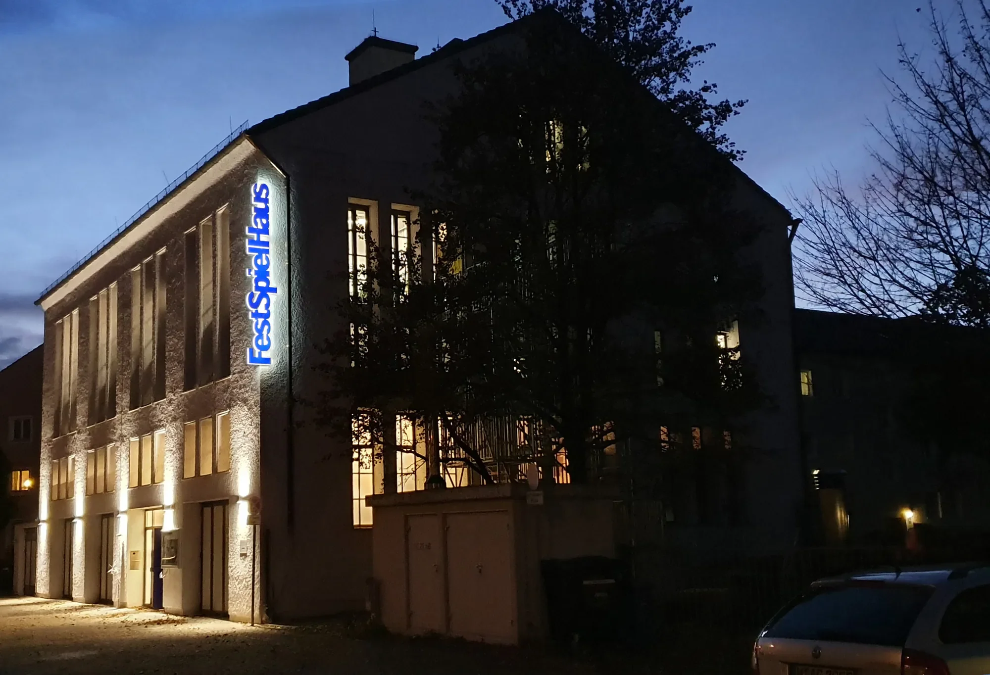 Ein dunkles Mehrfamilienhaus mit beleuchteten Fenstern und einem beleuchteten Firmenschild an der Seite, umgeben von Bäumen bei Dämmerung.