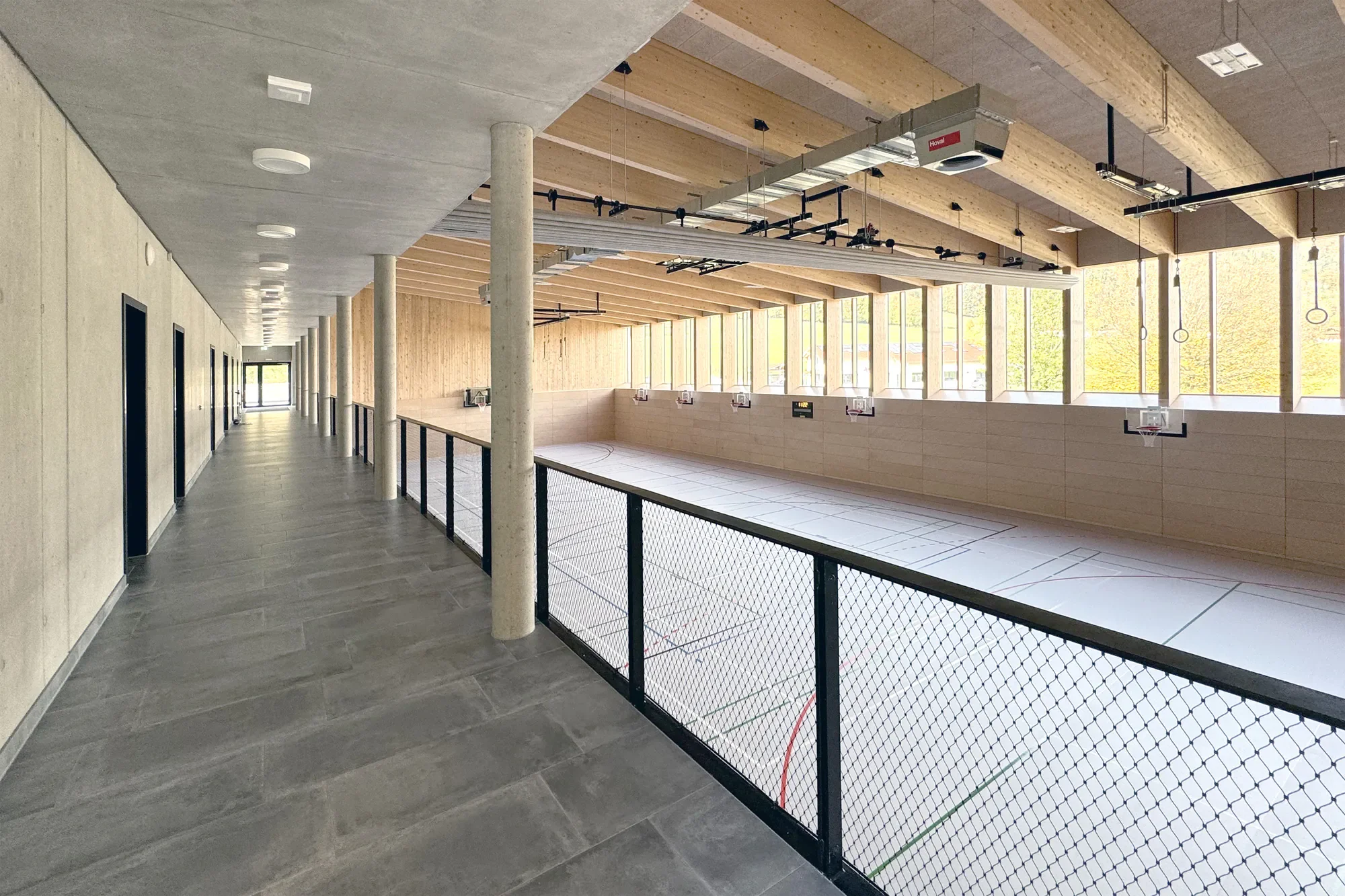 Innenansicht einer modernen Sportsporthalle mit Basketballplatz, große Fenster, Holzdecken, Flur auf der linken Seite mit Türen, Hallenboden für Basketballspiele, Netzabsperrung entlang des Spielfelds.