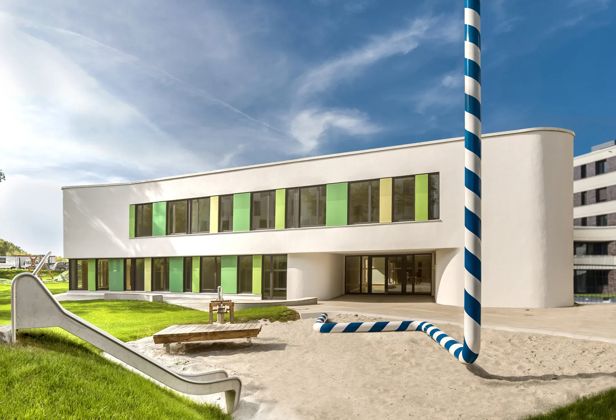 Modernes Gebäude mit bunten Fenstern und einem blauen und weißen Pfahl davor, auf einem Spielplatz mit Rutsche und Sand, bei sonnigem Himmel.
