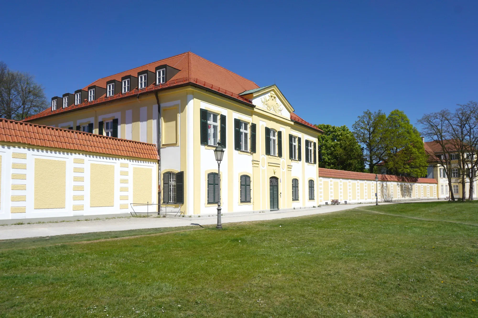 Ein gelbes Gebäude mit weißen Akzenten, schwarzen Fensterläden und rotem Ziegeldach, umgeben von grünen Bäumen unter einem klaren blauen Himmel.