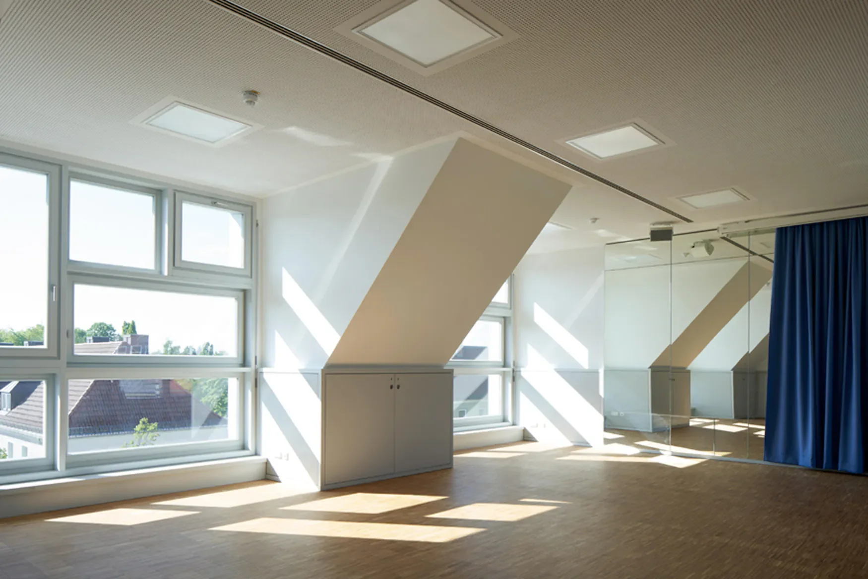 Ein heller Raum mit großen Fenstern, einer Spiegelwand und einer blauen Trennwand, Sonnenlicht strömt durch die Fenster, ausgestattet mit weißen Wänden, Holzfußboden und Deckenleuchten.