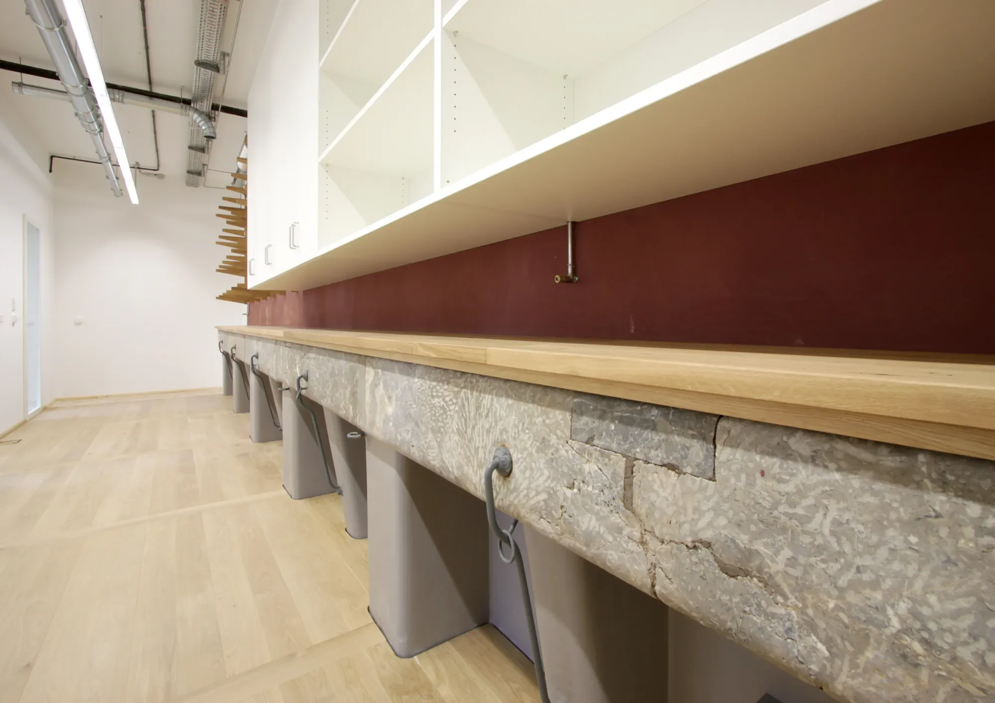 Ein Raum mit weißen Wänden, einer langen Theke mit einer Marmoroberfläche und offenen weißen Schrankfächern an der Wand. Über der Theke sind Haken an der Seite befestigt, die wahrscheinlich für Handtücher oder Schürzen gedacht sind. Im Hintergrund sind Lüftungsrohre und Kabel an der Decke sichtbar.
