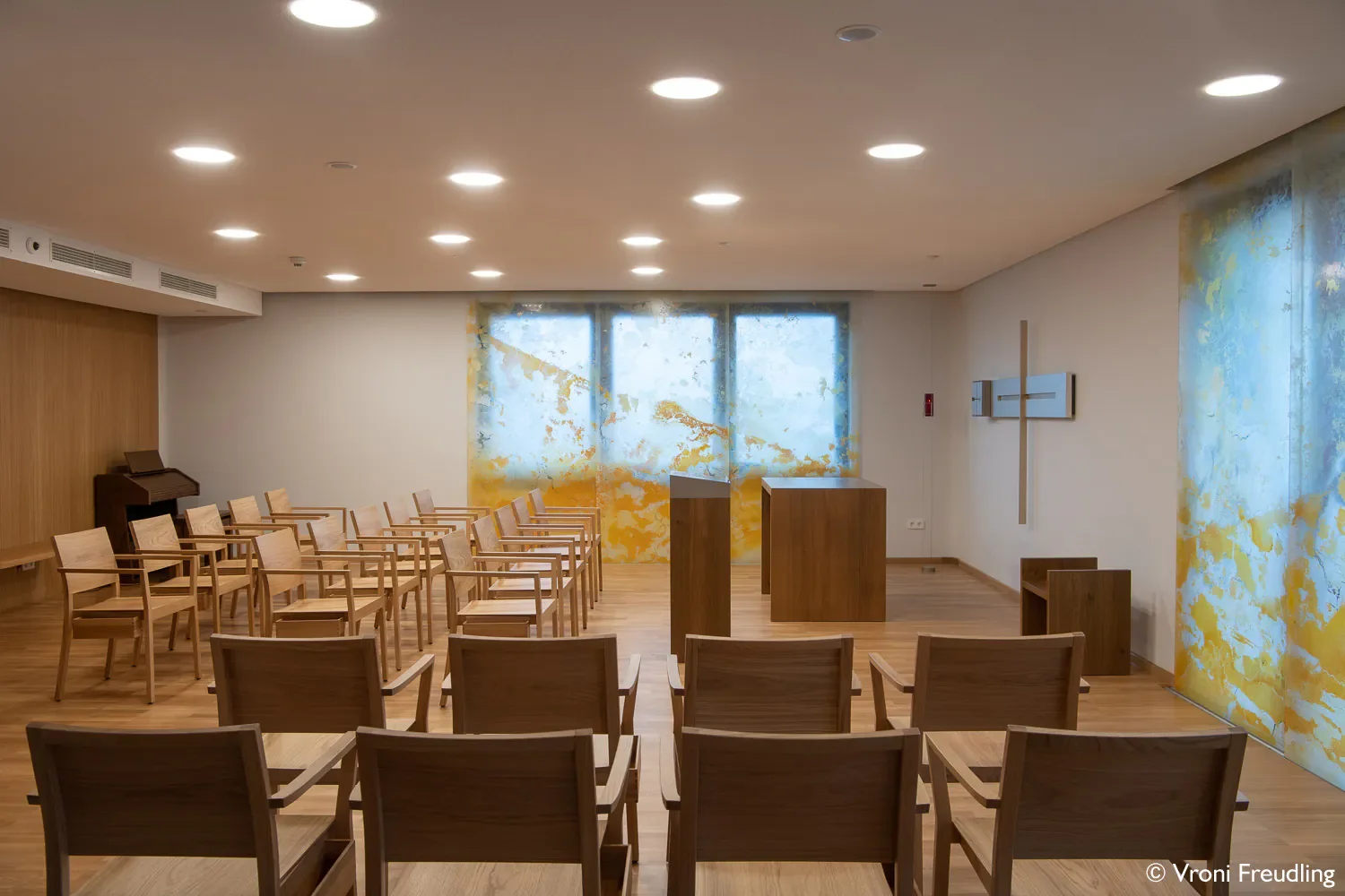 Ein leeres Kirchenraum mit Holzbänken, einer Orgel, einem Kreuz und bunten Wandbildern auf den Seitenwänden.