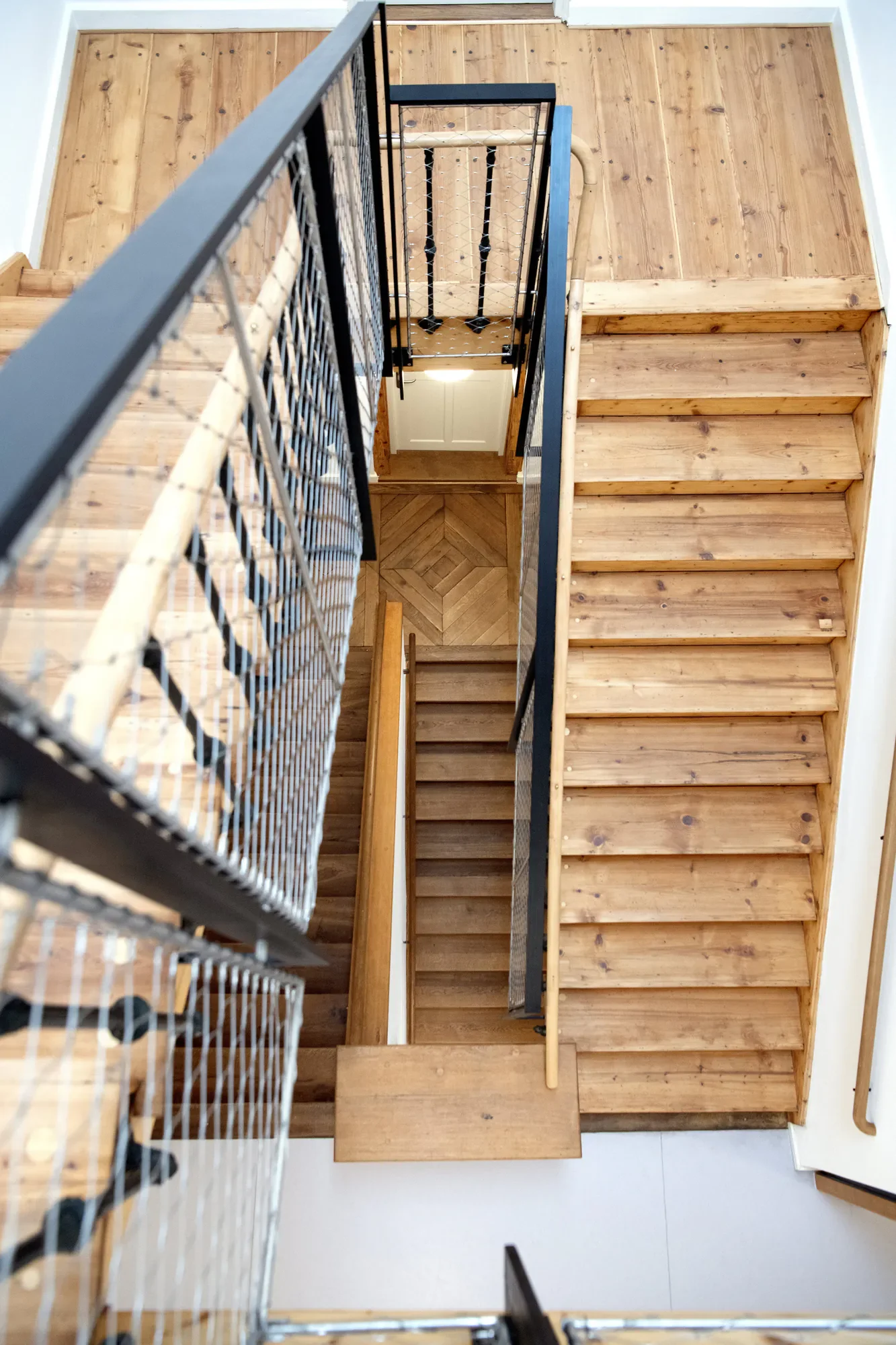 Ansicht eines Treppenhauses mit Holzstufen, Metallgeländer und Holzboden im oberen Stock. Die Perspektive ist nach oben gerichtet, mit einem Blick auf die Treppe und den Flur darunter.