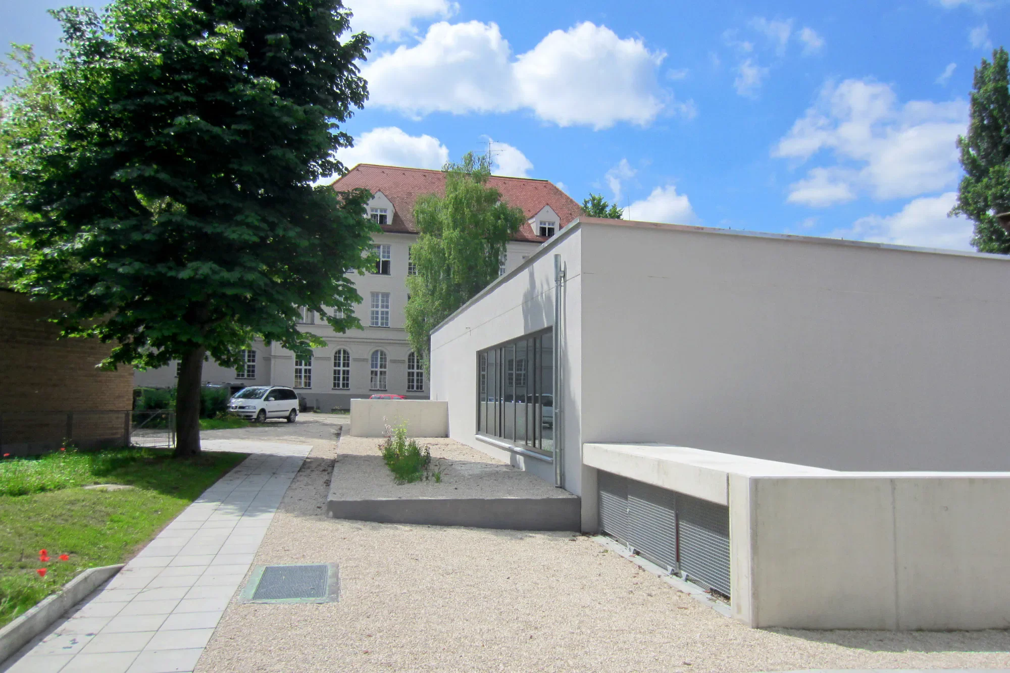 Moderne weiße Ecke mit großen Fenstern vor einer Gartenfläche, im Hintergrund alte Gebäude mit roten Dächern, Bäume und Himmel mit Wolken.