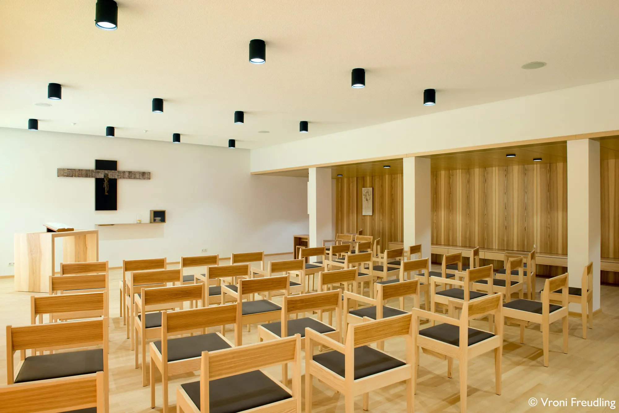 Innenraum eines Kirchsaals mit Stühlen, einer Wand mit Holzwandverkleidung und einem Kreuz an der Wand.