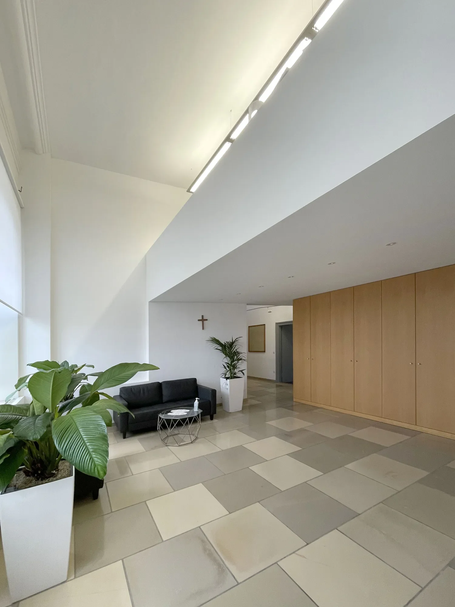 Ein Wartebereich in einem Gebäude mit grünen Pflanzen, schwarzen Sofas, einem kleinen Tisch und einem Holzschrank. Die Decke hat elektrische Beleuchtung und an der Wand ist ein Kruzifix zu sehen.