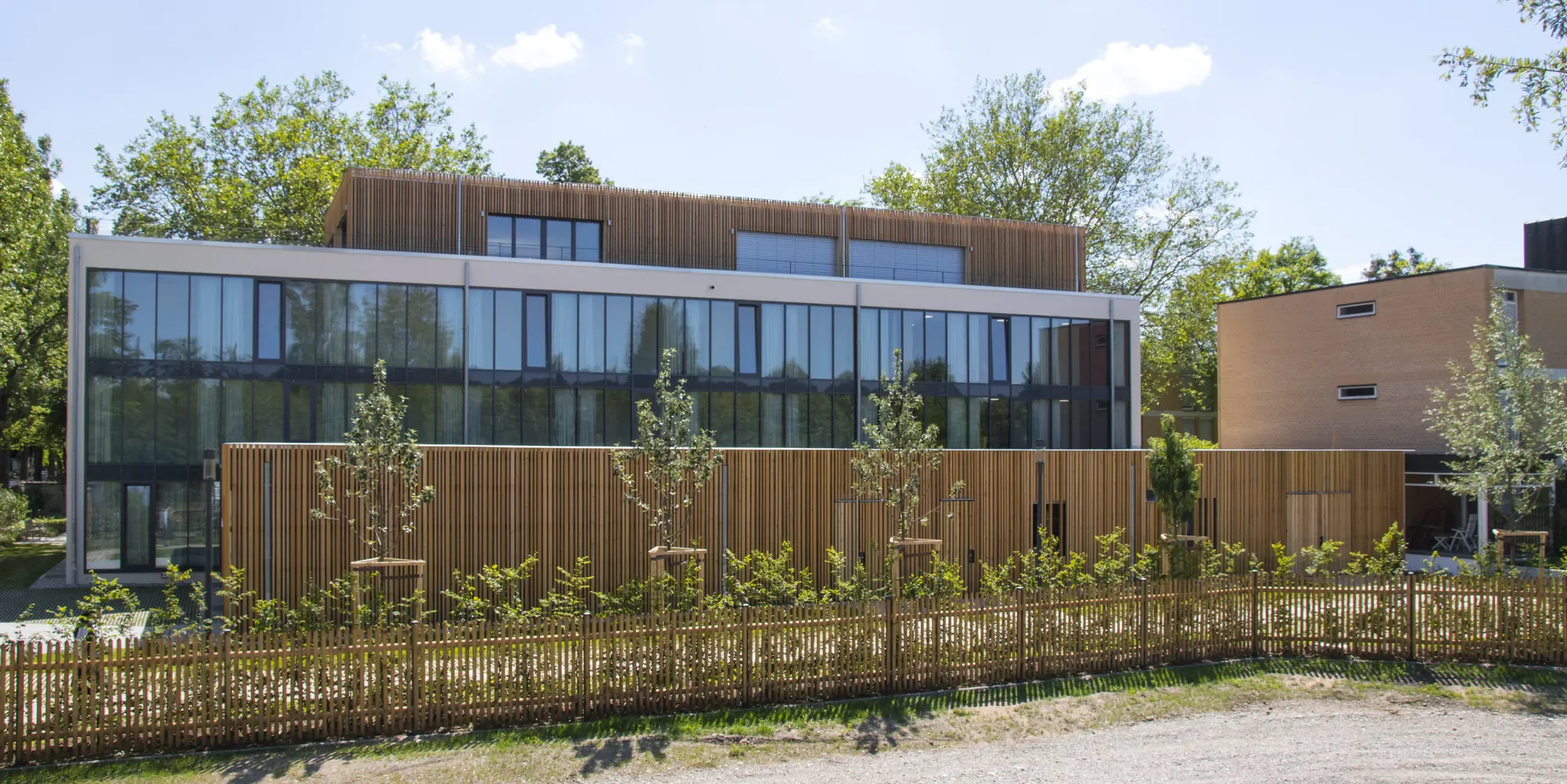 Modernes mehrstöckiges Gebäude mit großen Glasfenstern, Holzverkleidung und begrüntem Gelände im Vordergrund, umgeben von Bäumen und blauen Himmel.