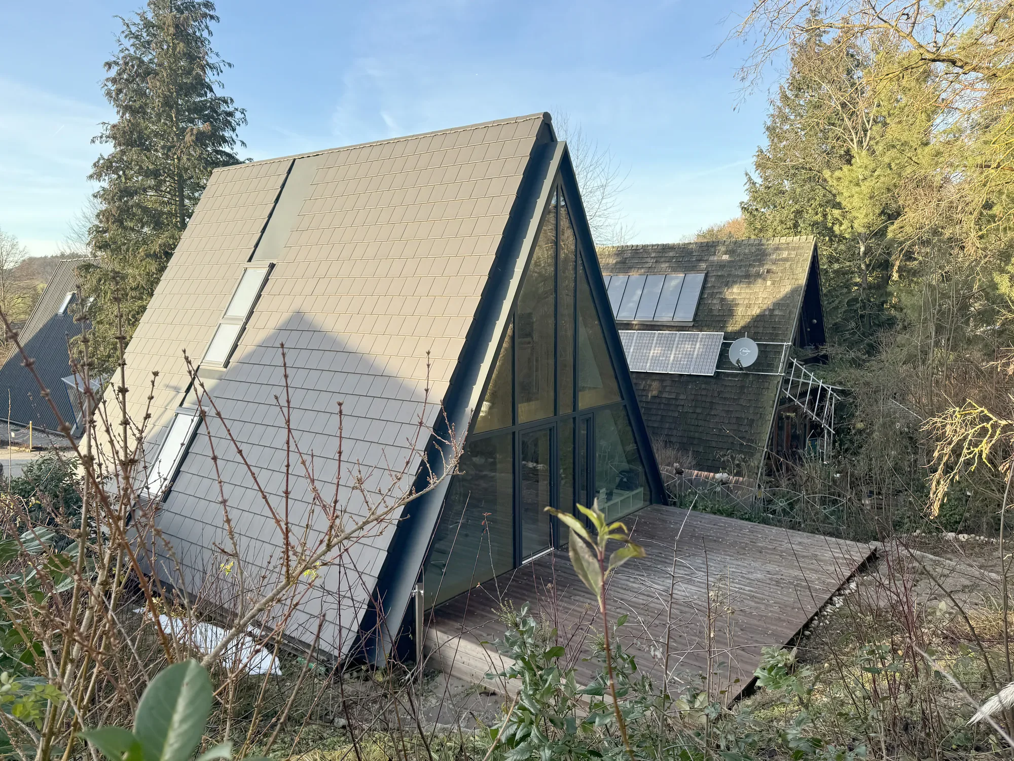 Modernes Haus mit großen Glasflächen und Solarpaneelen auf dem Dach, umgeben von Bäumen und Sträuchern, bei sonnigem Himmel.