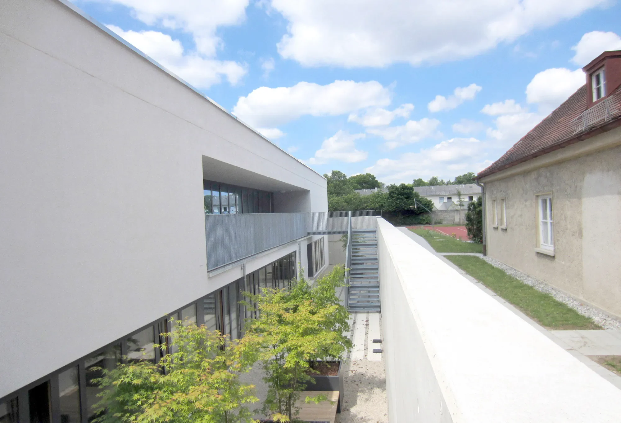 Ein moderner Gebäudekomplex mit weißer Fassade und großen Fenstern, umgeben von Bäumen und einem kleinen Garten, bei blauem Himmel mit Wolken.